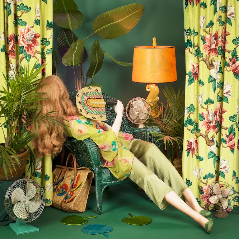 Fan Club by Patty Carroll is an archival pigment print, available in an edition of 15. This photograph features a mannequin reclining in a green chair, fanning herself while being surrounded by plants and fans. The paper size is 30 x 30 inches and