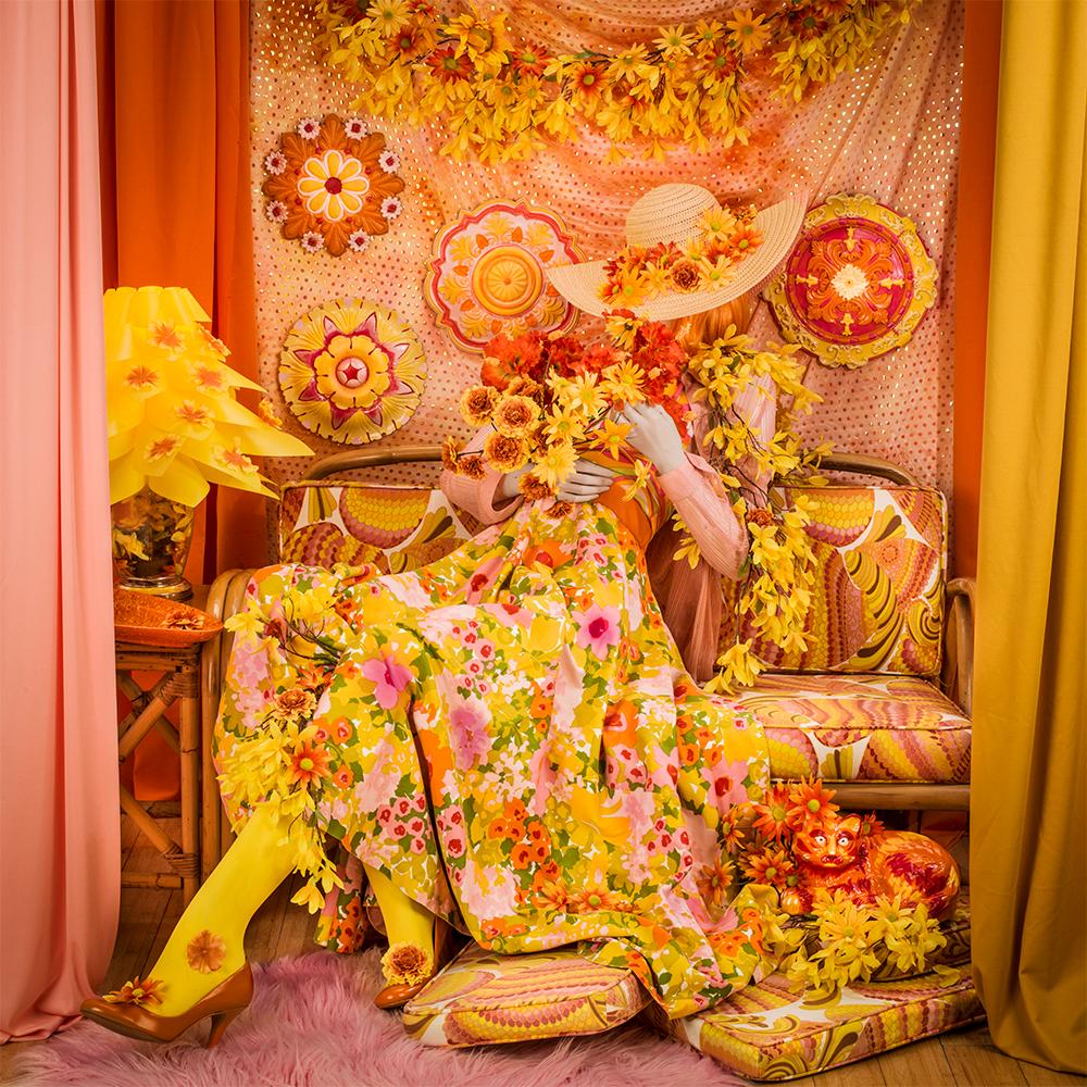 Flower Child by Patty Carroll presents vibrantly chaotic scene. A woman leans back in her couch, covered with yellow and orange flowers. Her floral patterned dress matches the color scheme of the room decor, the curtains, the couch, and the hanging