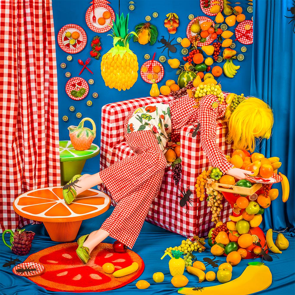 Fruity di Patty Carroll presenta una scena colorata e caotica che ricorda un picnic estivo. Una donna vestita con un plaid rosso è circondata da vari frutti e decorazioni a tema. Si confonde con la sedia a quadri, che imita una coperta da picnic,