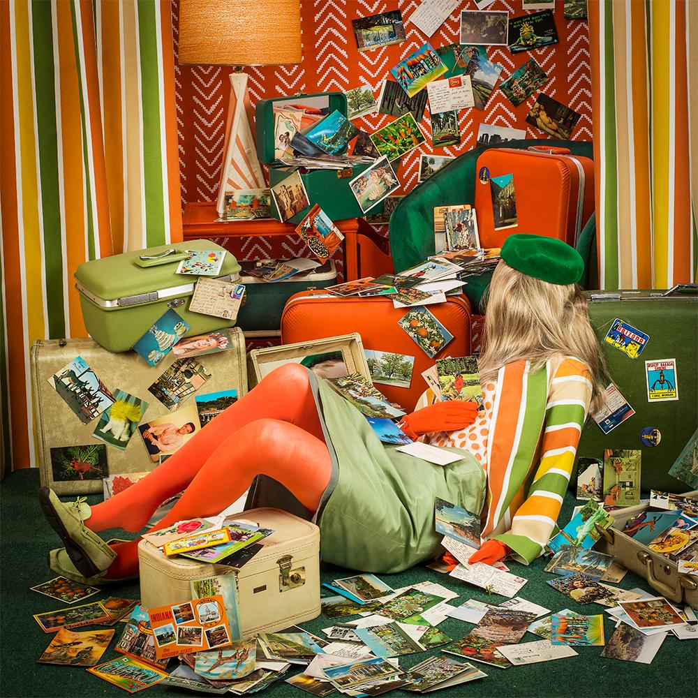 Gone Postal by Patty Carroll presents a chaotic scene, a room filled with orange, yellow and green luggage and scattered with postcards seeming to fall from above. A woman lies amidst the mess, leaning on the suitcases while she is covered in