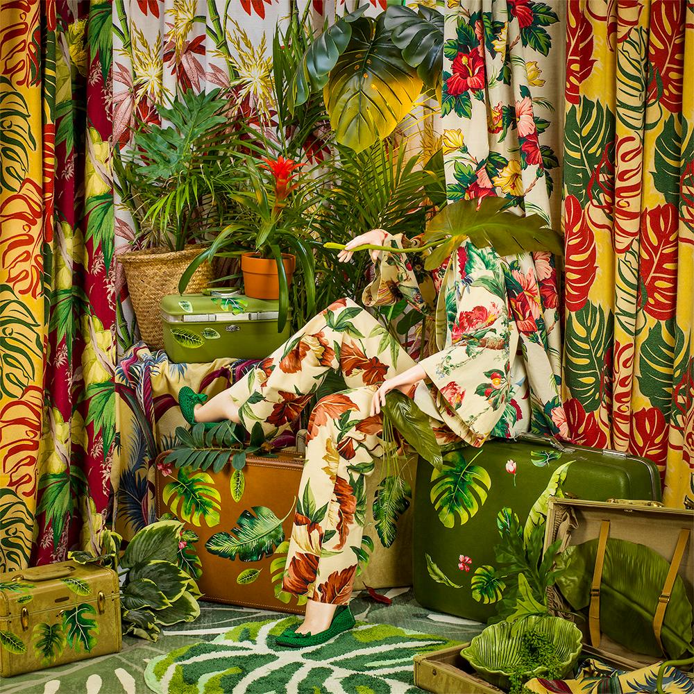 Leafing Home by Patty Carroll presents a chaotic botanical scene. A woman sits on top of suitcases covered in tropical leaves. She wears leaf printed clothing, and is surrounded by palm, monstera, and banana leaves. This theme is reflected in her
