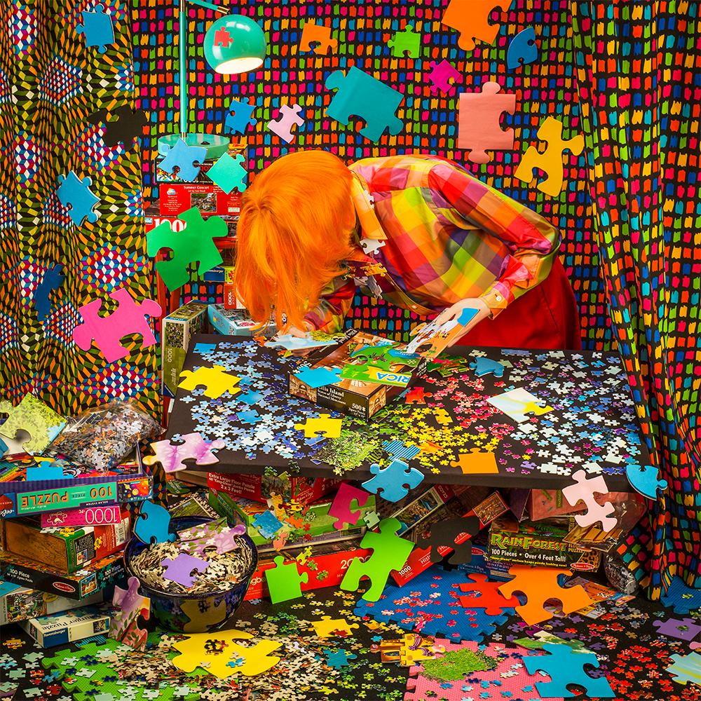 Puzzled de Patty Carroll présente une pièce chaotique aux couleurs vives. Une femme se tient penchée sur une table remplie de pièces de puzzle.  Des piles de boîtes de puzzle entourent la pièce, avec des pièces de puzzle se répandant sur le sol et