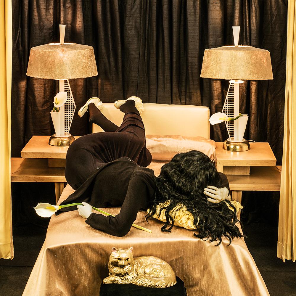 Rest of Her Life by Patty Carroll presents a luxurious gold and black room with gold decor. A woman lie backwards on the bed with her body twisted in a seemingly unnatural way. Calla lilies are found throughout the scene, one placed in each lamp and