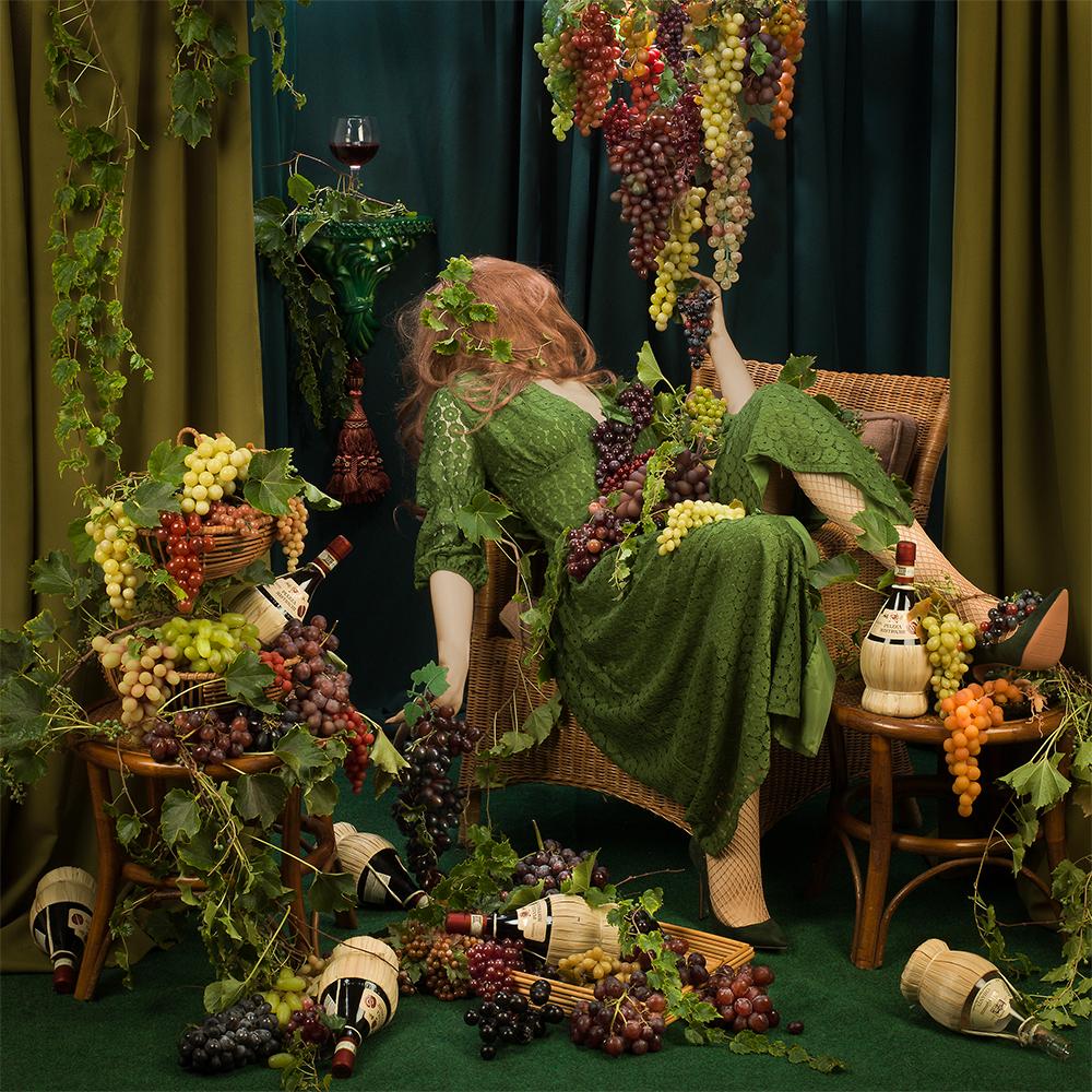 Sour Grapes by Patty Carroll presents a chaotic scene, a room filled by grapes, vines and wine bottles. A woman lies back in her chair, covered in the grapes and vines overtaking the room. 

This photograph's image size is 22 x 22 inches, with the