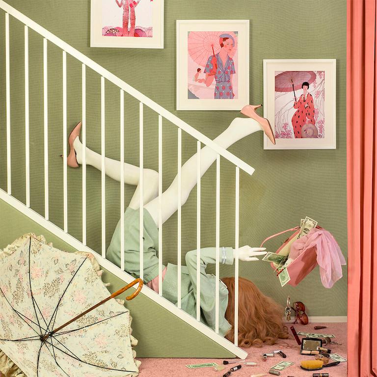 Staired Down by Patty Carroll presents a chaotic scene. A woman tumbles down the stairs, with her arms and legs flailing. Her purse spills out above her head, covering the floor with various beauty products and money. Soft green and pink tones fill