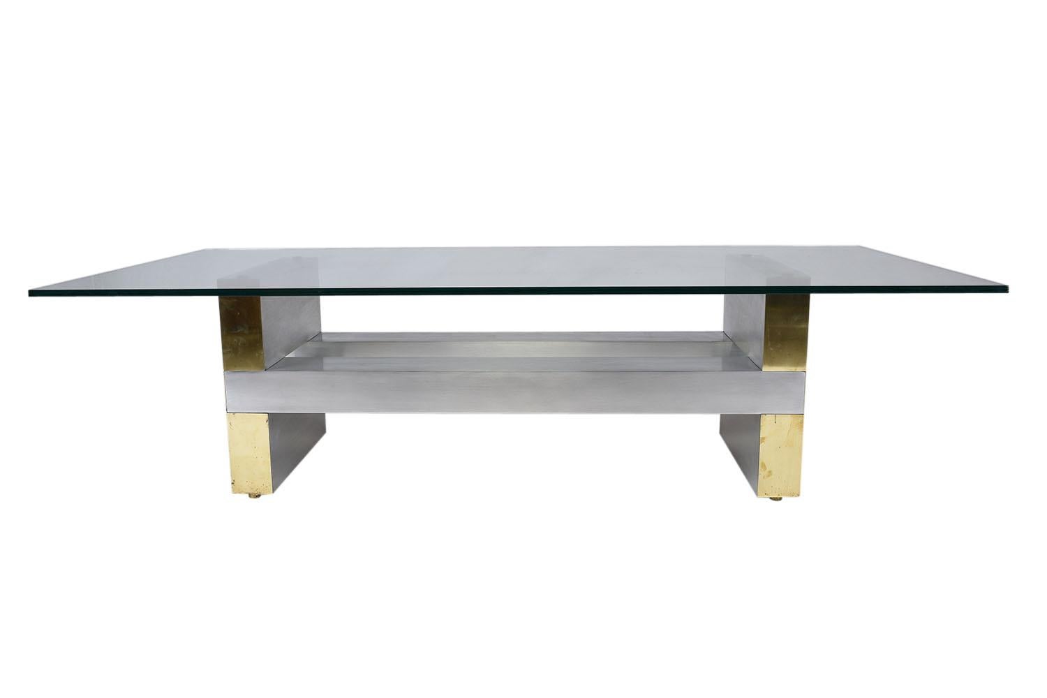 This 1960s Mid-Century Modern-style coffee table is in the manner of Paul Evans and features a geometric brushed steel and brass base. The clear glass tabletop is rectangular and features a flat polish. This vintage coffee table is sturdy, stylish,