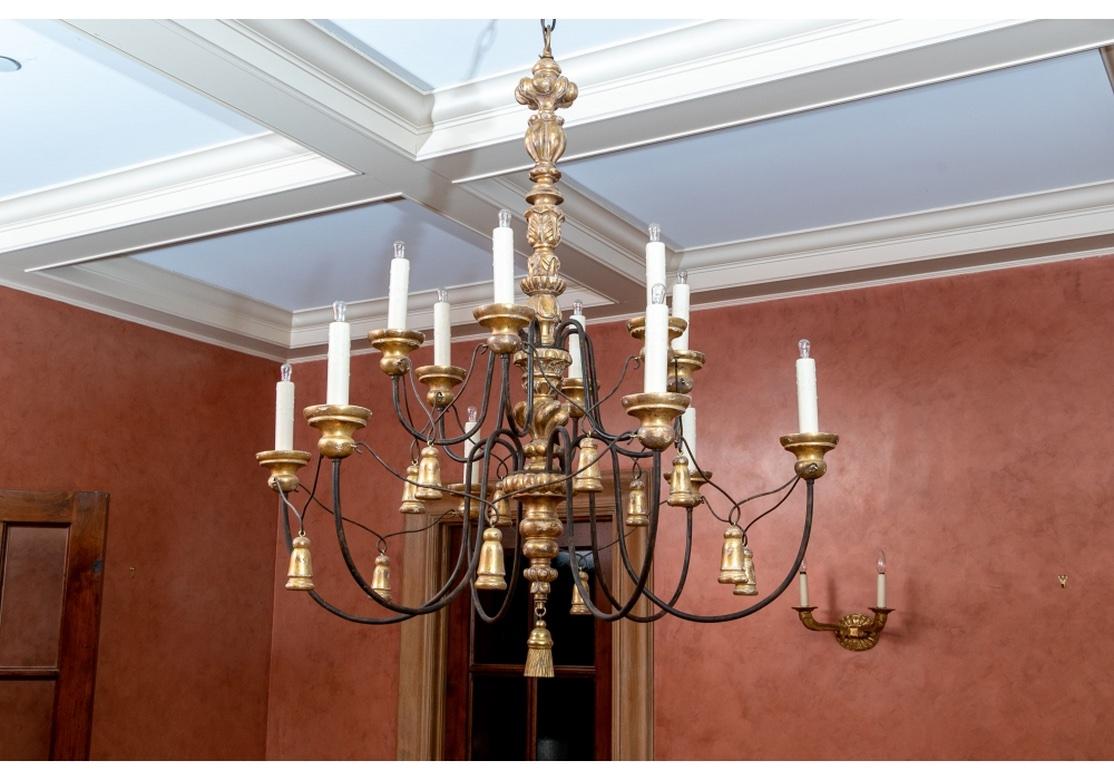 Large iron and carved gold leaf wood chandelier with 16 candlelights and the arms conjoined with draping tassel motifs. White paper candle covers with faux drip. A carved leafy and knobbed gilt standard and fluted ceiling plate, with a