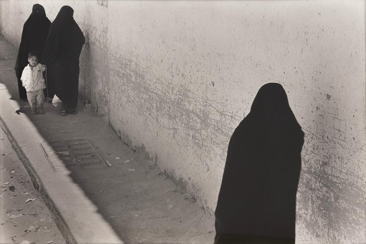 Three Figures in Black - Morocco by Paul Greenberg depicts four people walking on a sidewalk. A small child walks with two figures cloaked in black, and carries a white toy attached to a string. Another lone figure stands on the opposite side. It's