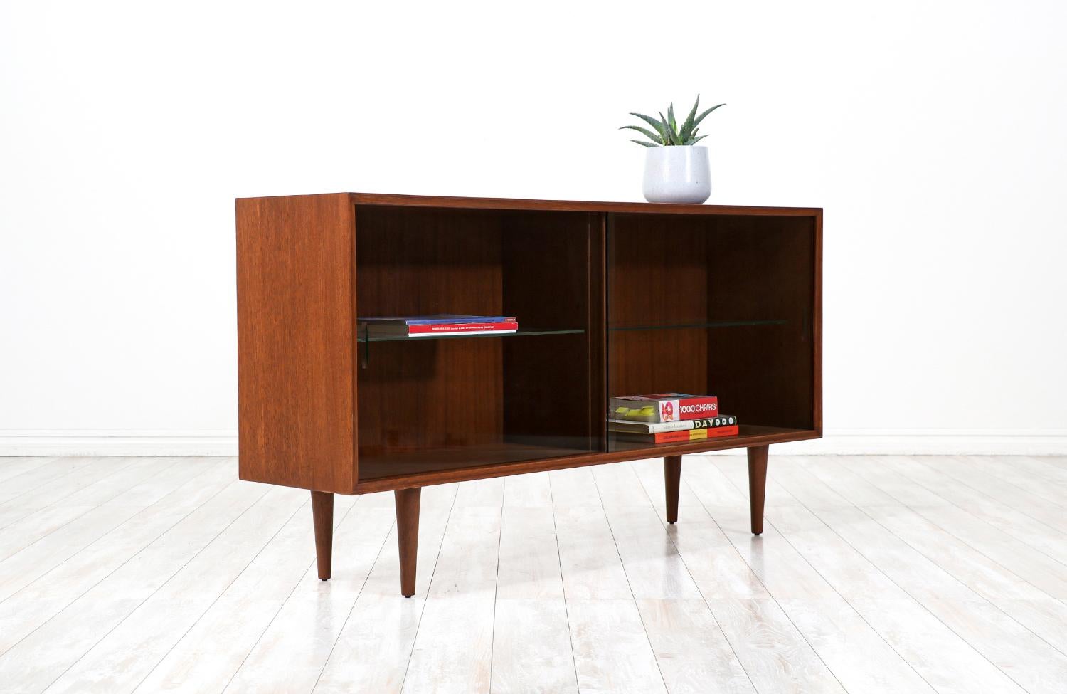 Mid-Century modern bookcase designed by Paul Laszlo and manufactured for Brown Saltman in United States circa 1950s. This iconic design features a walnut-stained mahogany case with a glass shelved interior, with glass doors, and two beautiful