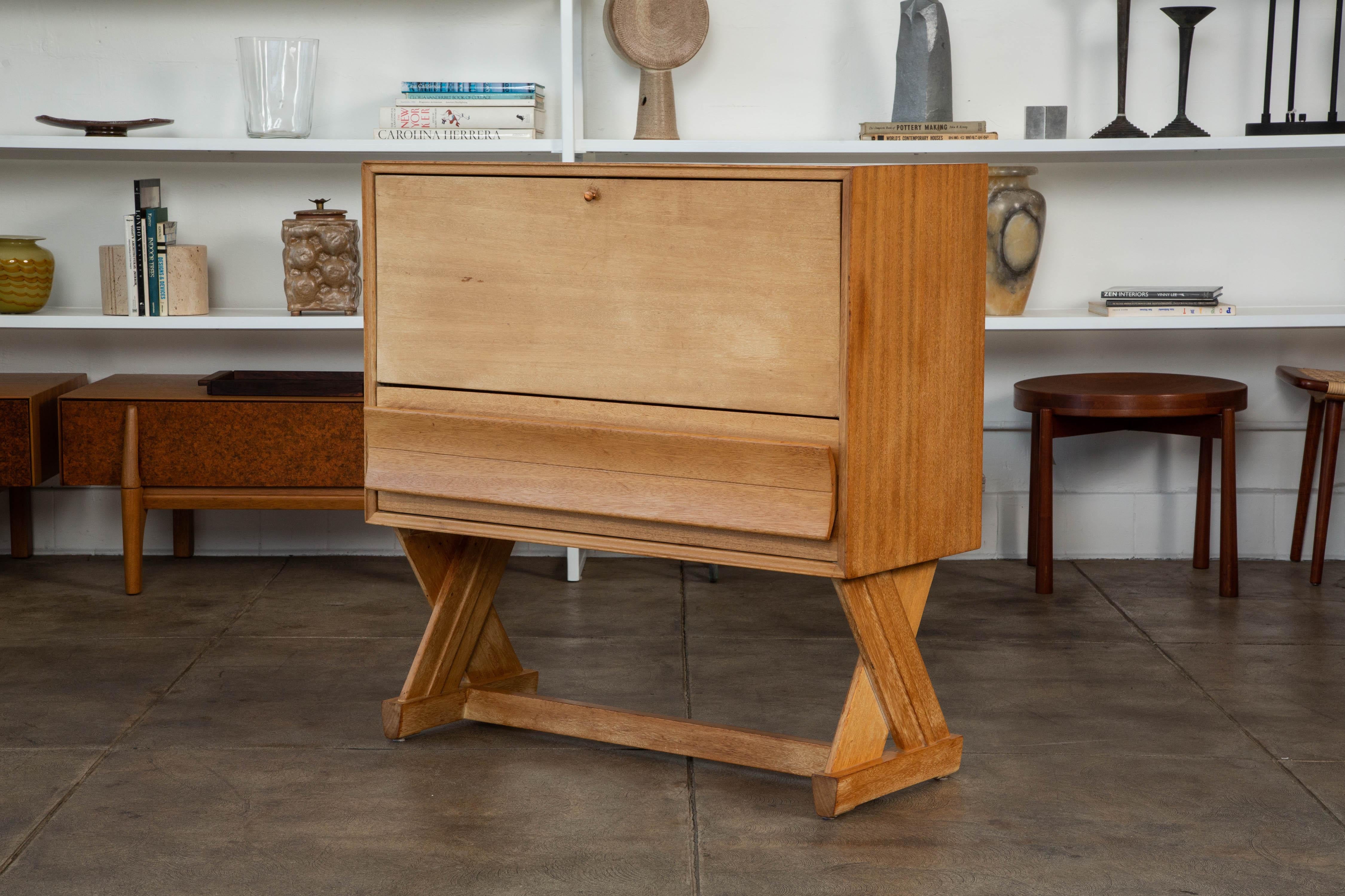 Liquor bar by Paul Laszlo for Brown Saltman, c.1957. This liquor cabinet features a bleached mahogany body and X-shaped legs with a pull down front. The top portion features vertical separation and thin lower storage drawer. The front panel drops