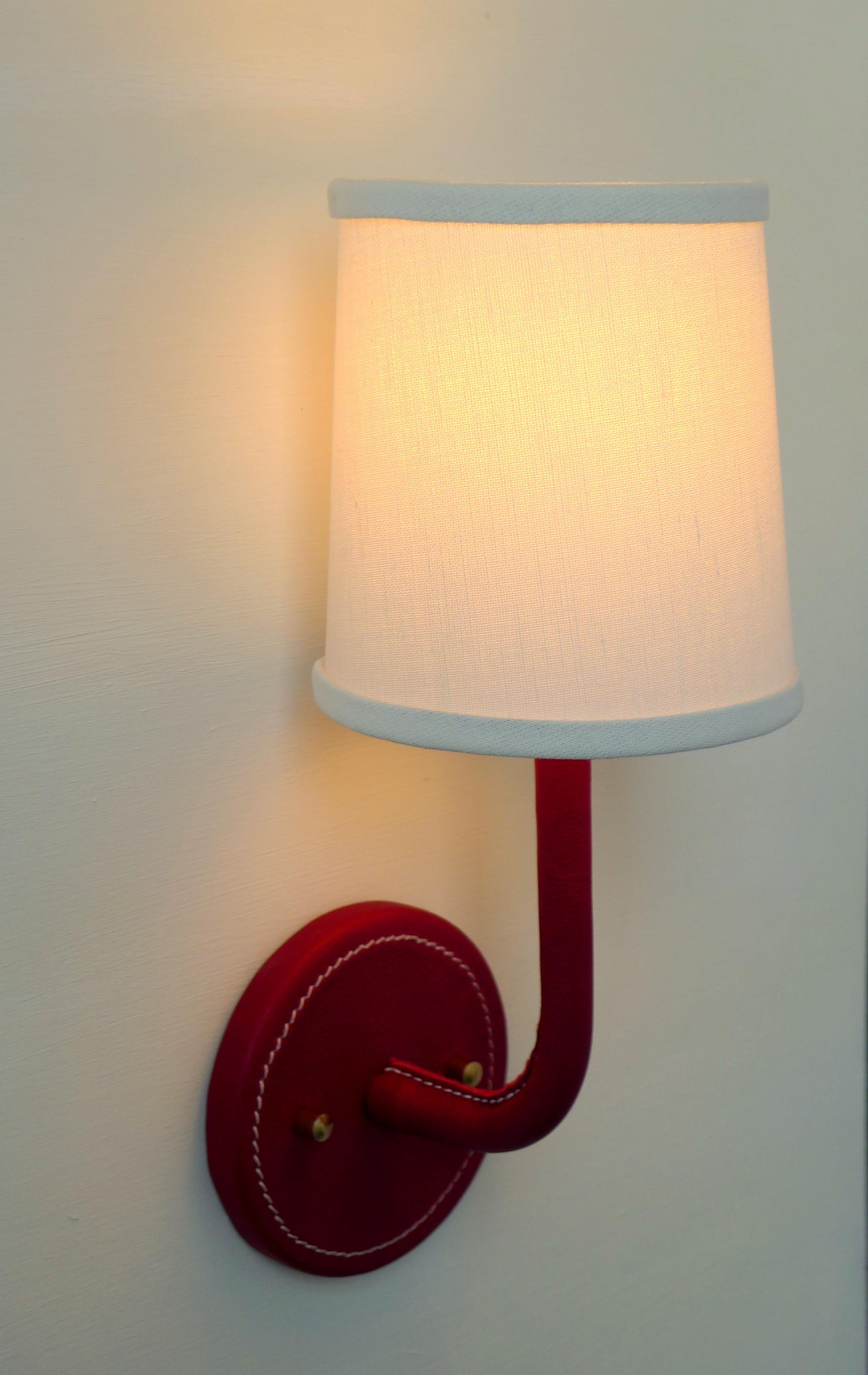 Paul Marra red leather wrapped Adnet inspired wall sconce, with top-stitched detailing. Top-stitching detail is on back plate and inner arm. Linen shade. Brass hardware. Dimensions provided are overall with shade. The shade is 4.5 top and 5.5