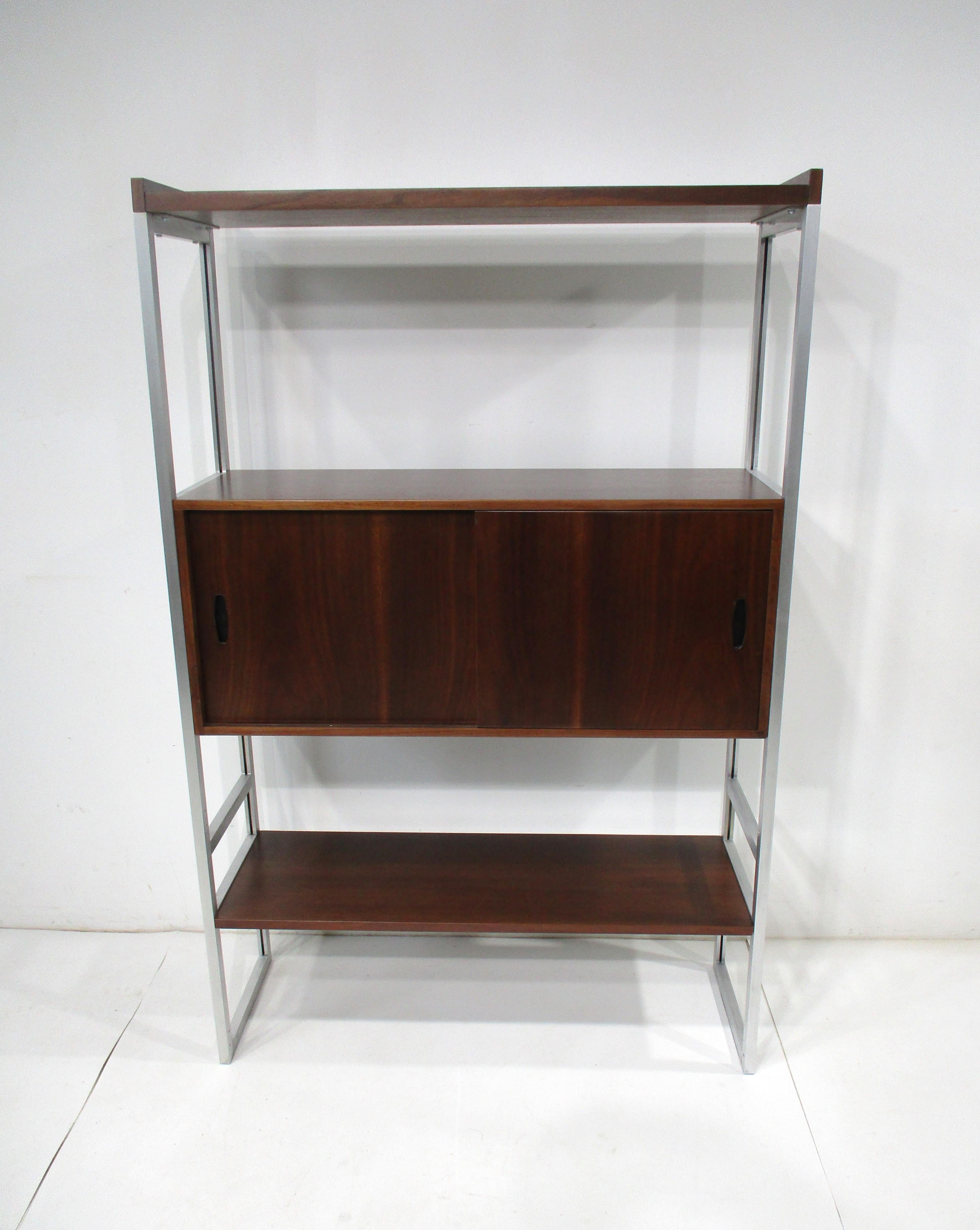A Mid Century walnut bookcase with two shelves and a small cabinet with two sliding doors for storage . Sitting on a aluminum frame and free standing this smaller scaled piece would fit into any tight spaces , interiors or office and with the