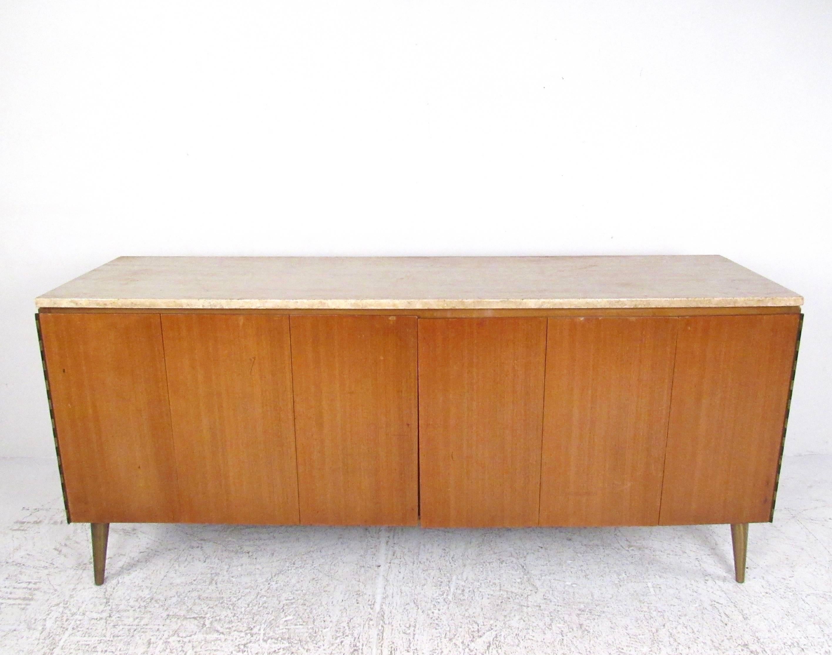 This stylish midcentury sideboard features the iconic design of Paul McCobb and boasts vintage mahogany finish and a striking travertine top. Tapered brass legs add to the appeal of this American Modern credenza designed for Calvin Group. Please