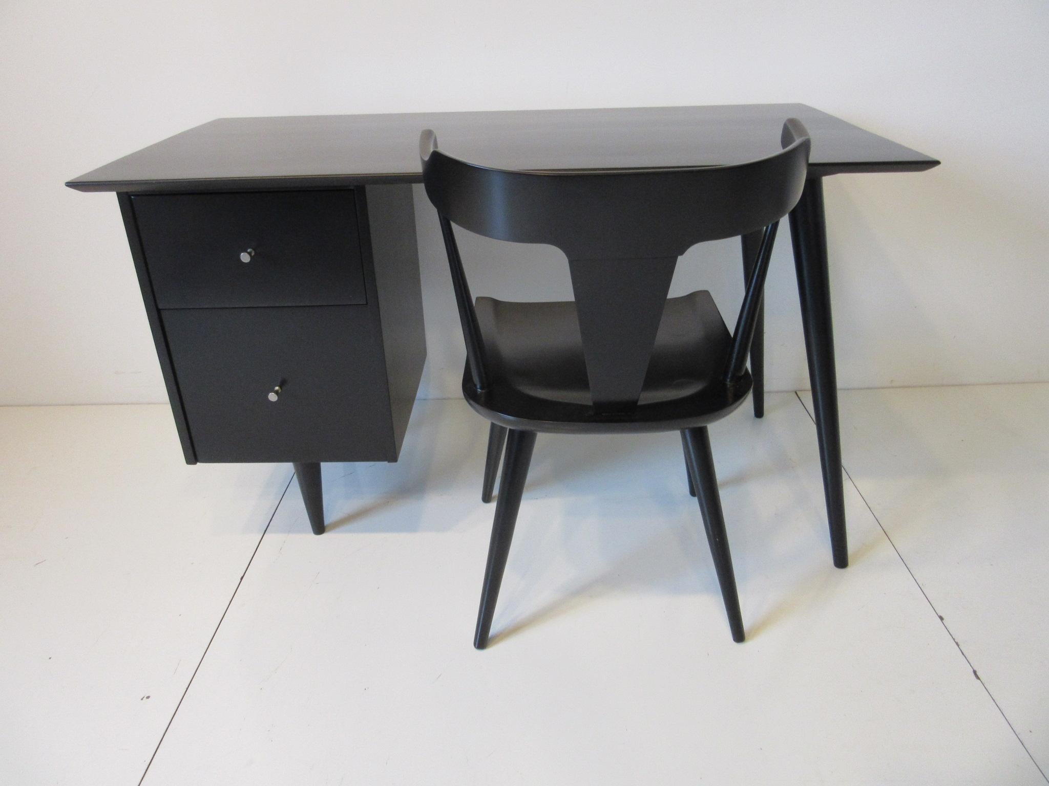 A desk and matching T- backed chair designed by Paul McCobb in solid maple with a satin black finish having brass pulls. Two drawers one a file size make this a very usable desk for working at home, stylish with two conical legs to the other side.