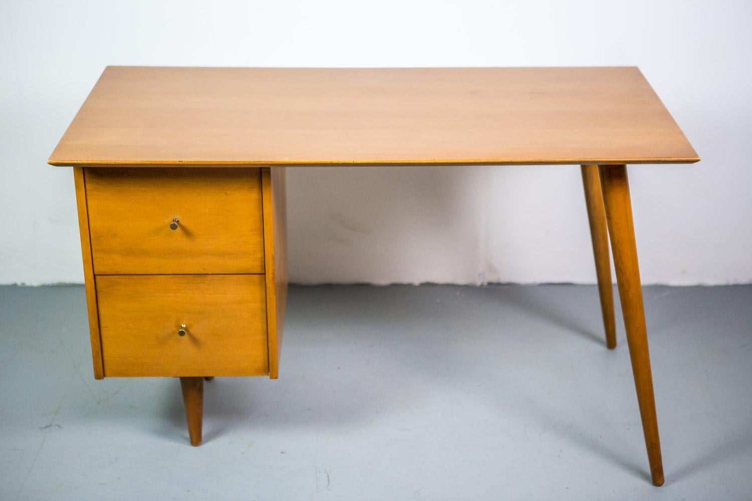 This exceptional early desk was designed by Paul McCobb and produced by Planner Group. Executed in solid maple, angular tapered legs and two desk drawers with conical brass pulls.