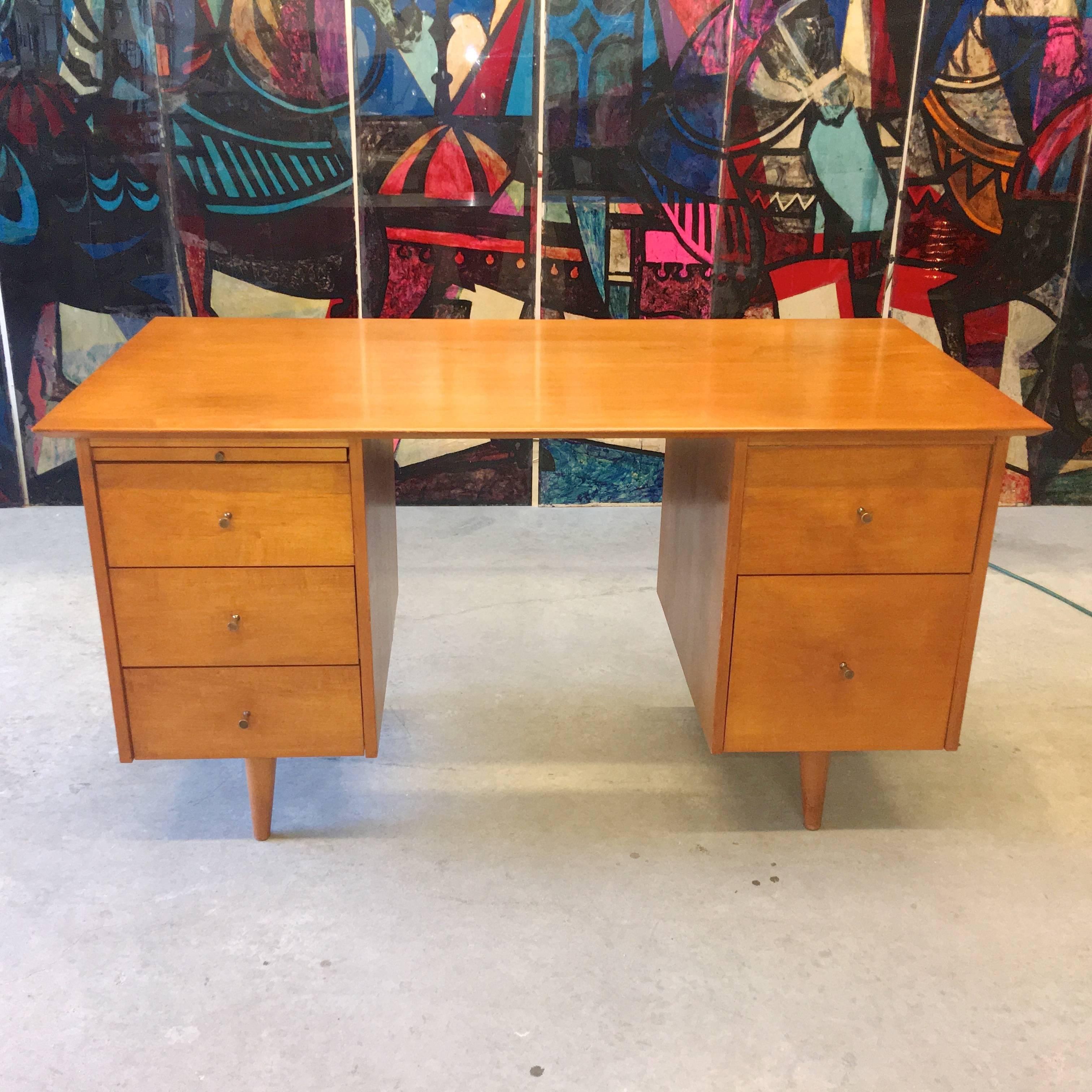 This iconic desk designed in 1950 by Paul McCobb for Winchendon Furniture Co. has a Classic design constructed of solid maple in original natural finish with five drawers and a pull-out work surface and distinctive brass conical pulls. Kneehole 20