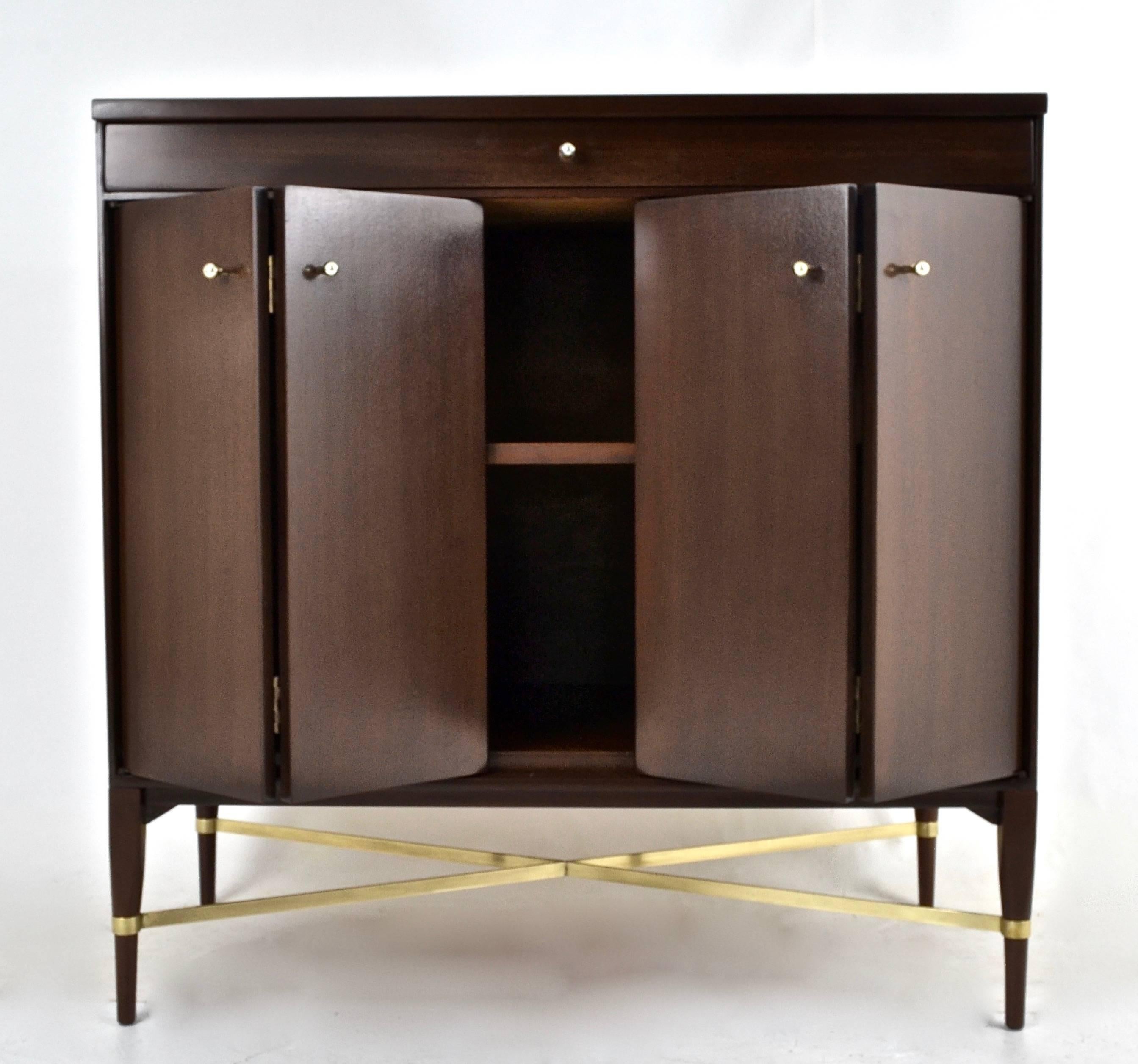 Featuring signature brass cross stretcher, this unusual McCobb cabinet has been fully restored. Single long drawer over a pair of bi-fold doors. Original pulls. Brass stretcher has been polished and lacquered.