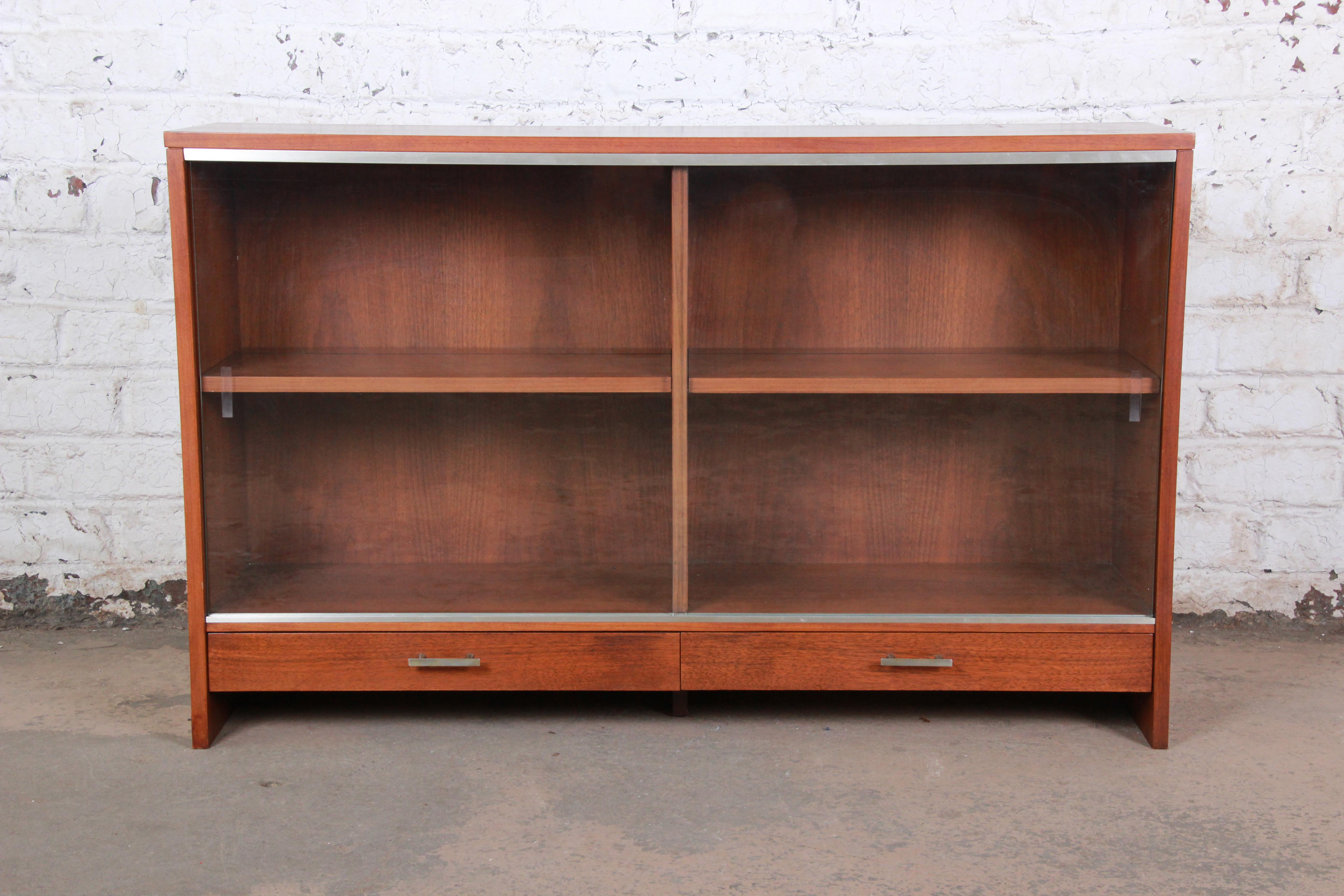 A sleek and stylish Mid-Century Modern glass front display cabinet or bookcase designed by Paul McCobb for Calvin furniture. The bookcase features beautiful walnut wood grain, with aluminum trim and hardware. It offers good storage, with two
