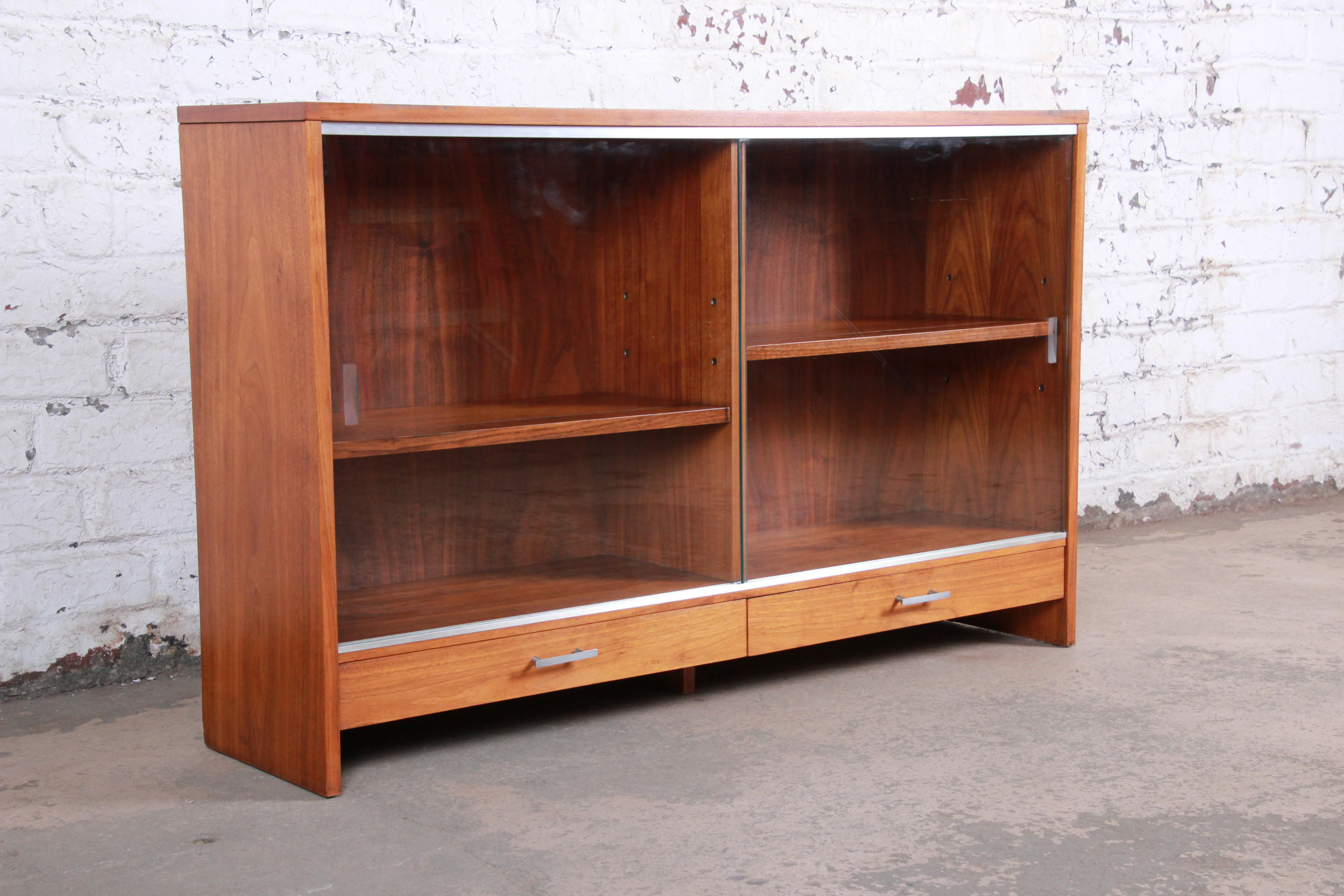 A sleek and stylish Mid-Century Modern glass front display cabinet or bookcase designed by Paul McCobb for Calvin Furniture. The bookcase features beautiful walnut wood grain, with aluminum trim and hardware. It offers good storage, with two