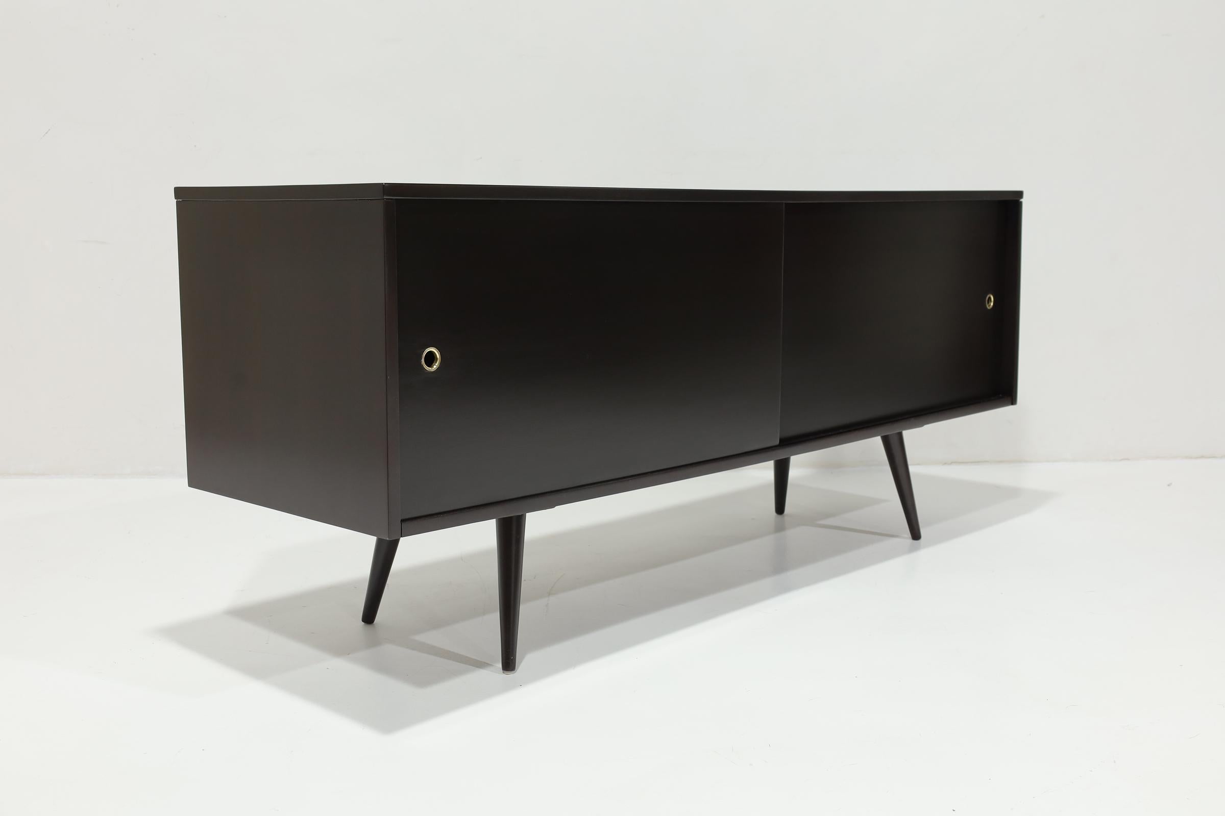 Sleek midcentury credenza/sideboard by Paul McCobb for Planner Group, 1950s. Stained in an espresso-colored stain. The sideboard has brass pulls and a single adjustable shelf on each side. 