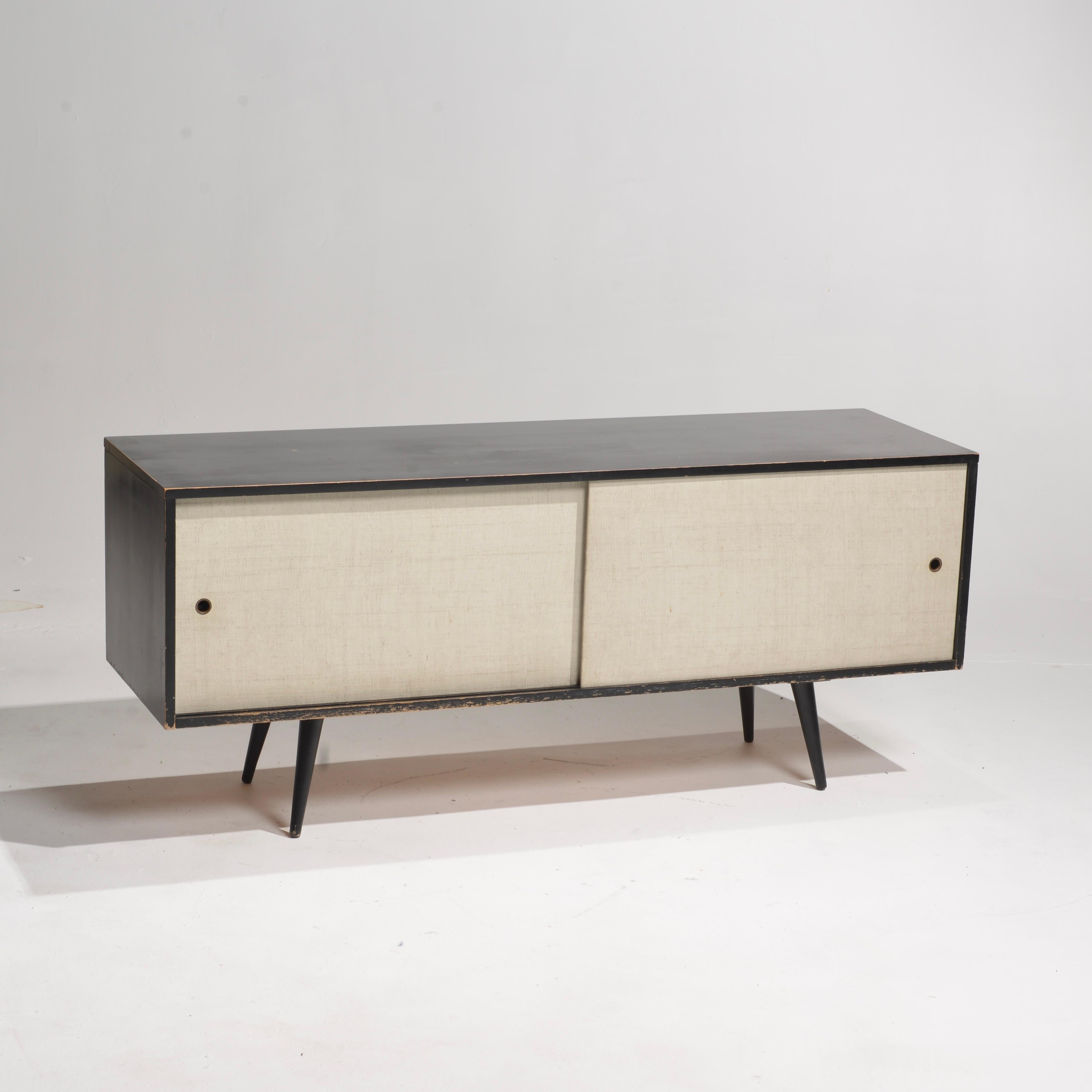 Bellissima credenza bassa di metà secolo di Paul McCobb, circa 1950, Planner Group. 1 scaffale regolabile con perni. Struttura in acero massiccio con finitura originale in lacca nera. Le ante scorrevoli in stoffa bianca con tiretto in ottone