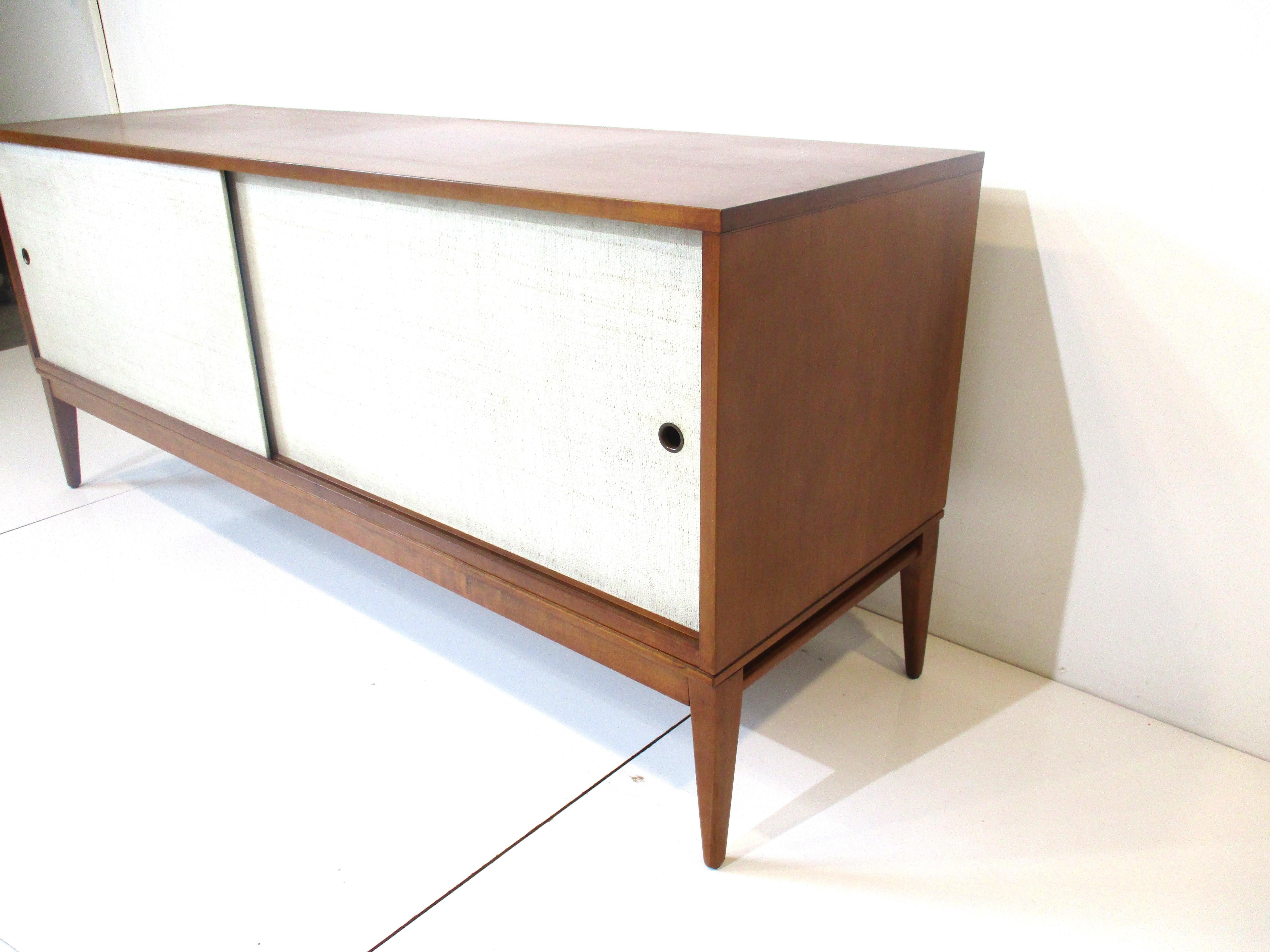 A low profile two sliding door credenza in a medium walnut toned finish with creamy beige door fronts. Metal finger holes for pulls and tapered legs to each corner, still retains the foil manufactures label to the backside designed by Paul McCobb