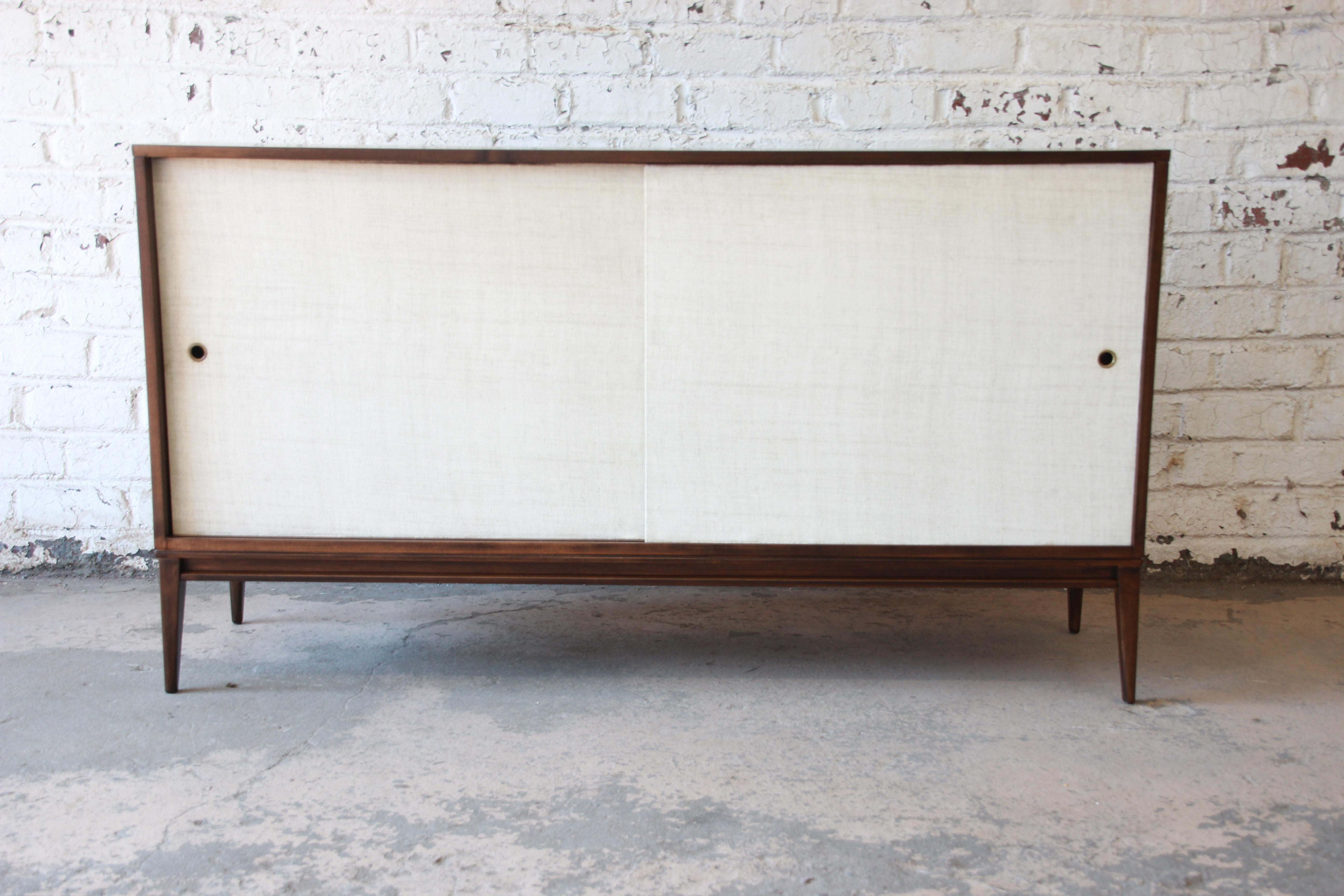 An exceptional Mid-Century Modern sideboard credenza from the iconic Planner Group line by Paul McCobb for Winchendon Furniture. The sideboard is made from solid birch and has two white grasscloth sliding cabinet doors. The left door opens up to a