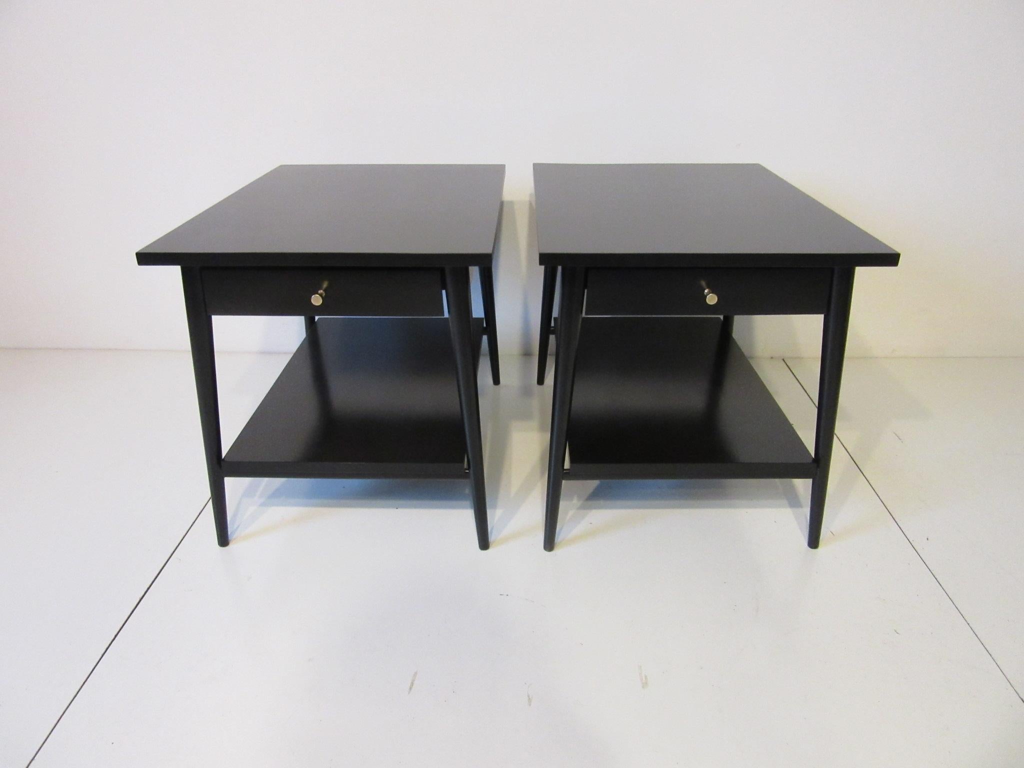 A pair of satin black nightstands or end tables with single drawer, solid brass T pulls and lower shelve . They retain the manufactures label to each piece crafted by the Winchendon Furniture company and from the Planner Group Collection.