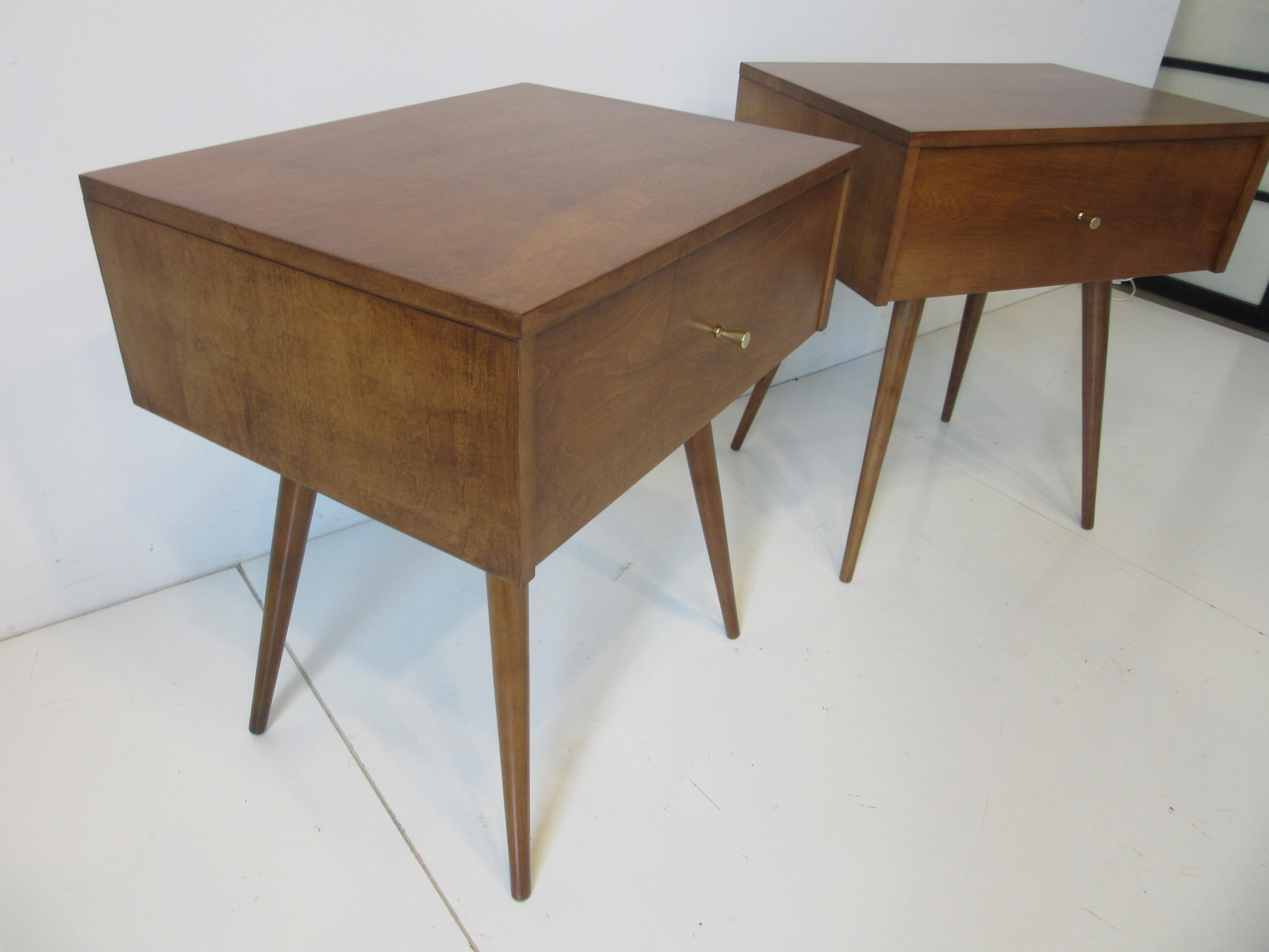 A pair of mid century medium walnut toned nightstands sized for today's higher pillow top matrasses with conical legs, each having one drawer with solid brass golf tee styled pulls . A great simple light look and feel crafted in solid wood typical