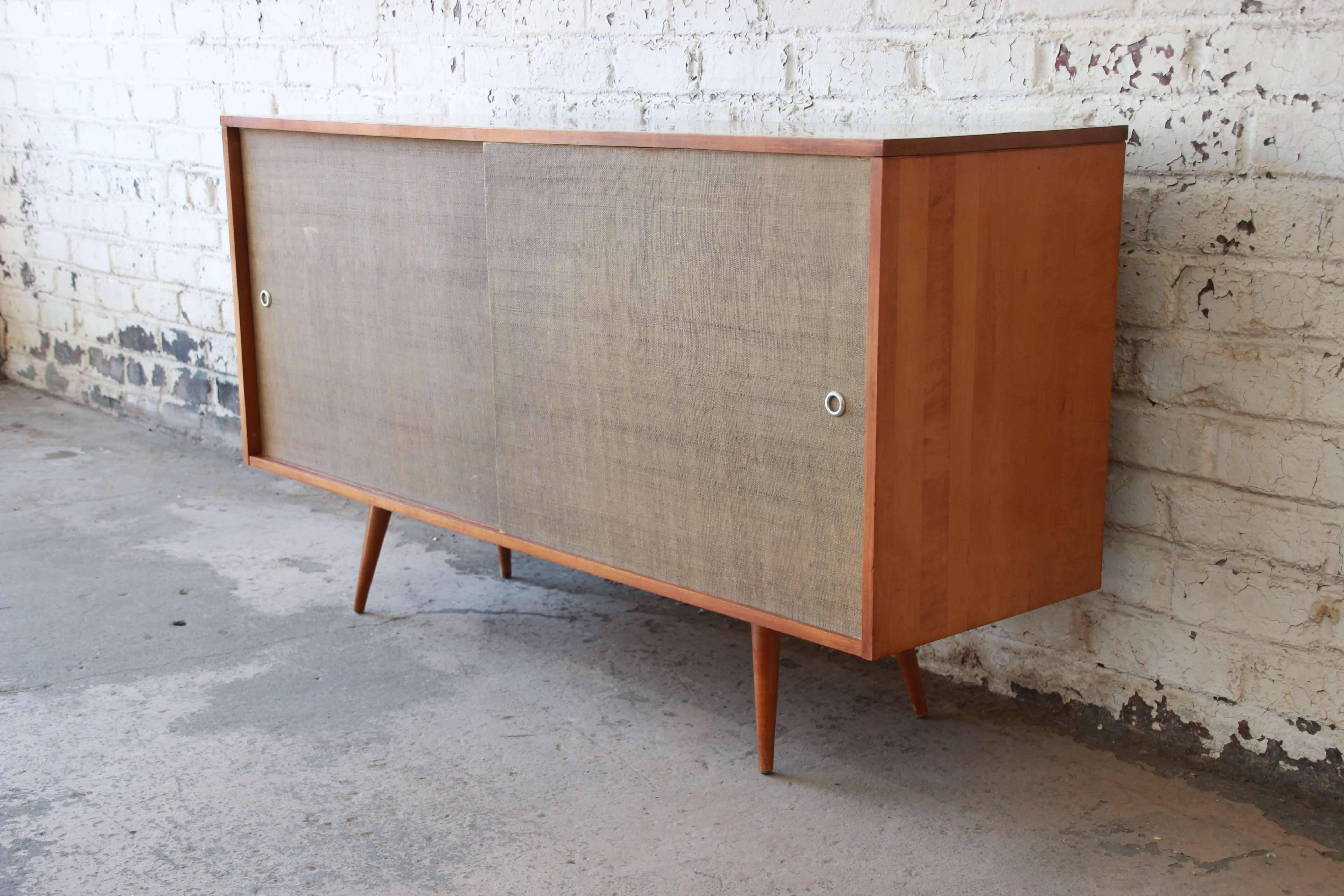 Offering a nice original planner group Paul McCobb sideboard credenza for Winchendon Furniture. The piece is made from solid birch and has two grass cloth front sliding cabinet doors. The left door opens up to a drawer and adjustable shelf while the