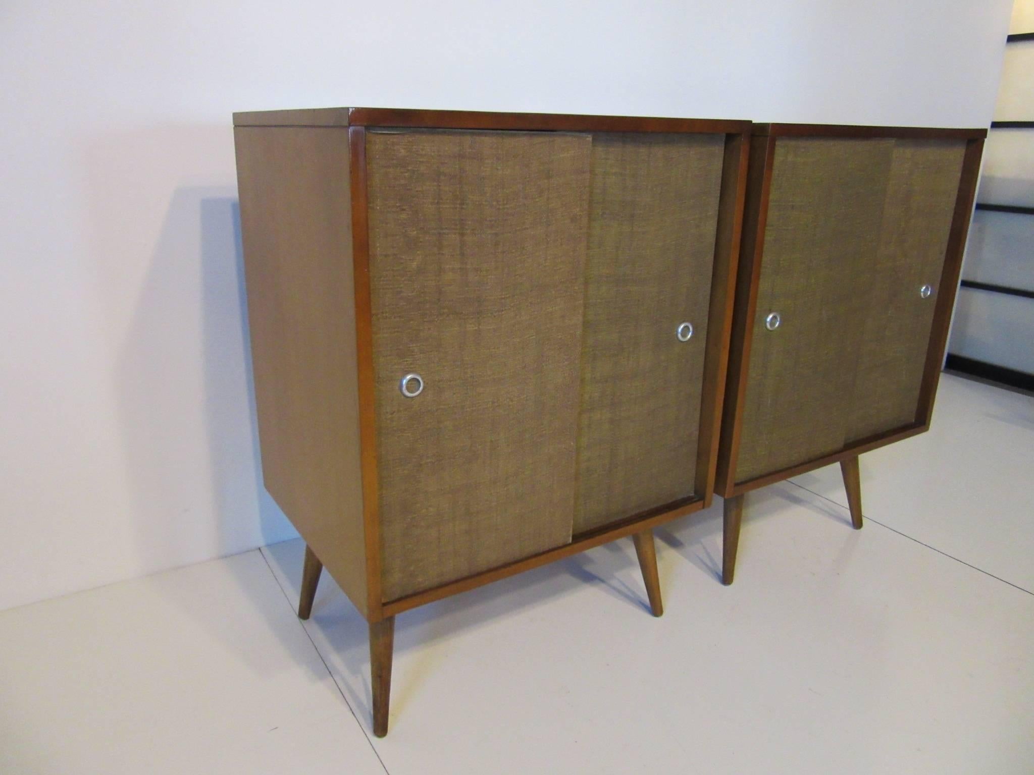 A pair of sliding grass cloth door smaller sized cabinets with fixed conical legs manufactured by the Winchendon Furniture Company for the Planner Group Collection.