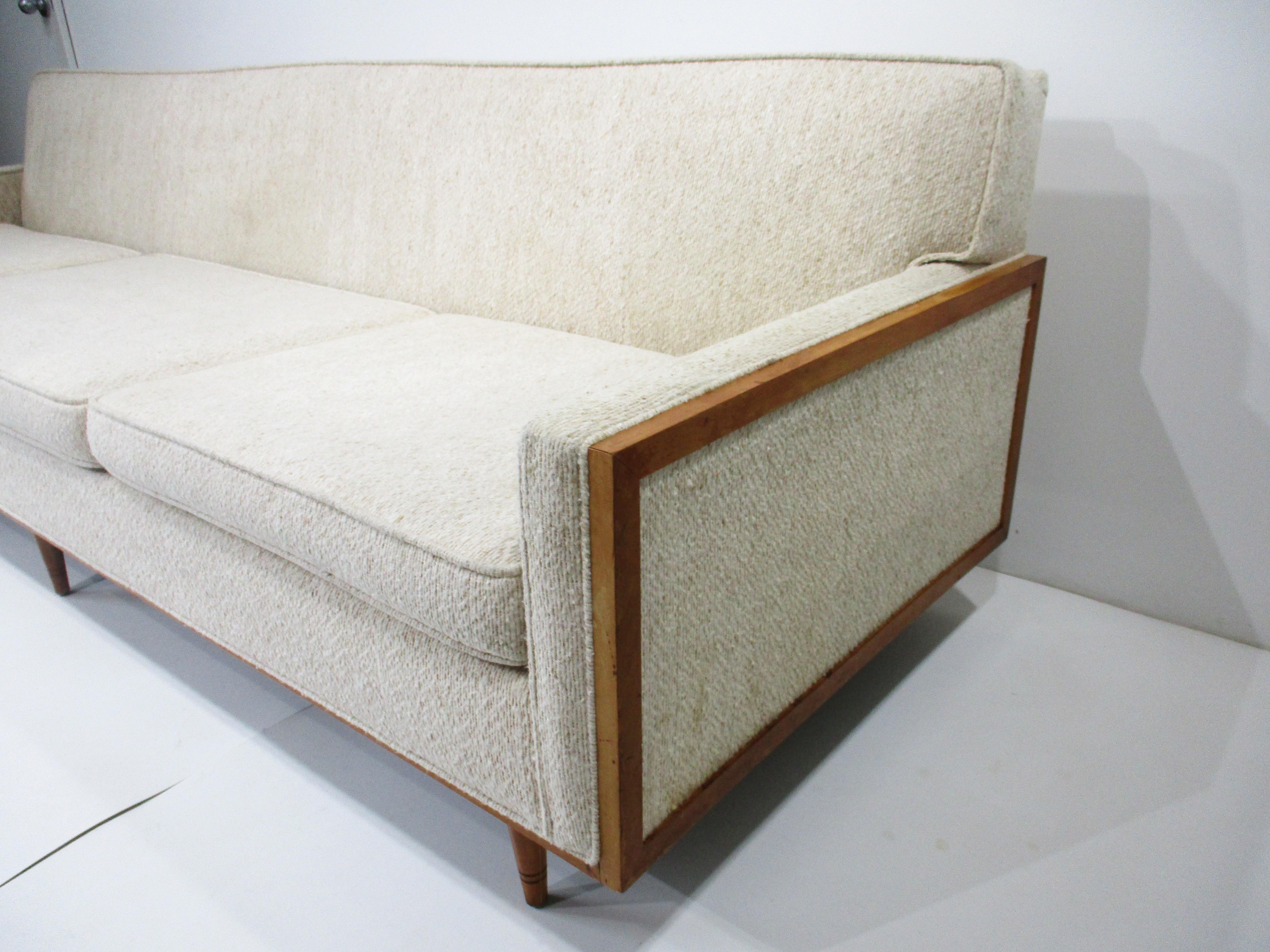 A Mid Century sofa with very clean lines having birch wood accents to each arm end and lower front , sitting on conical legs . Designed in the manner of Paul McCobb and upholstered in a woven lighter beige toned styled chenille fabric , a