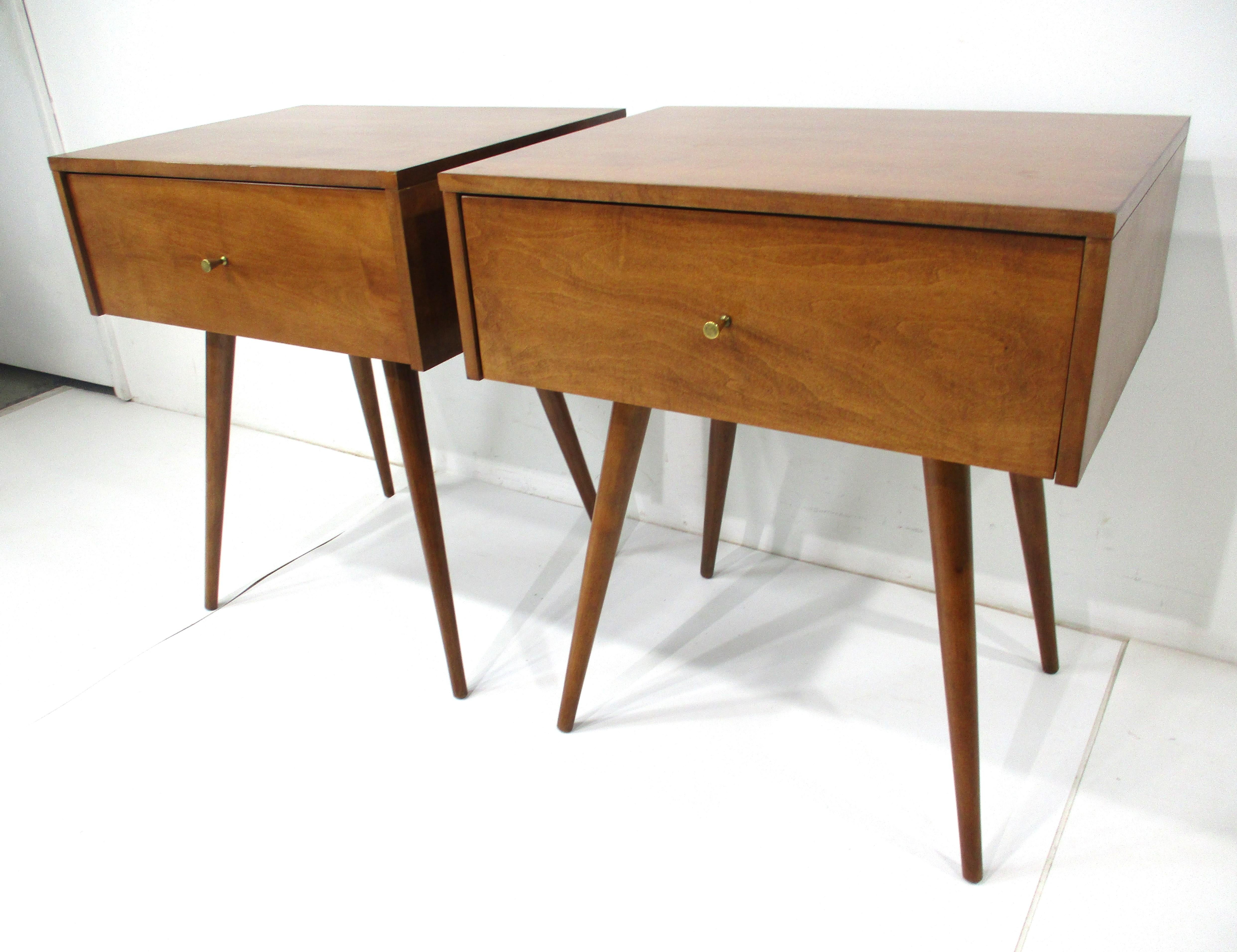 A pair of medium walnut toned solid maple wood nightstands with a single drawer to each having brass pulls . Siting on conical legs which are in a higher profile to match todays mattresses heights with a form that is simple but functional able to