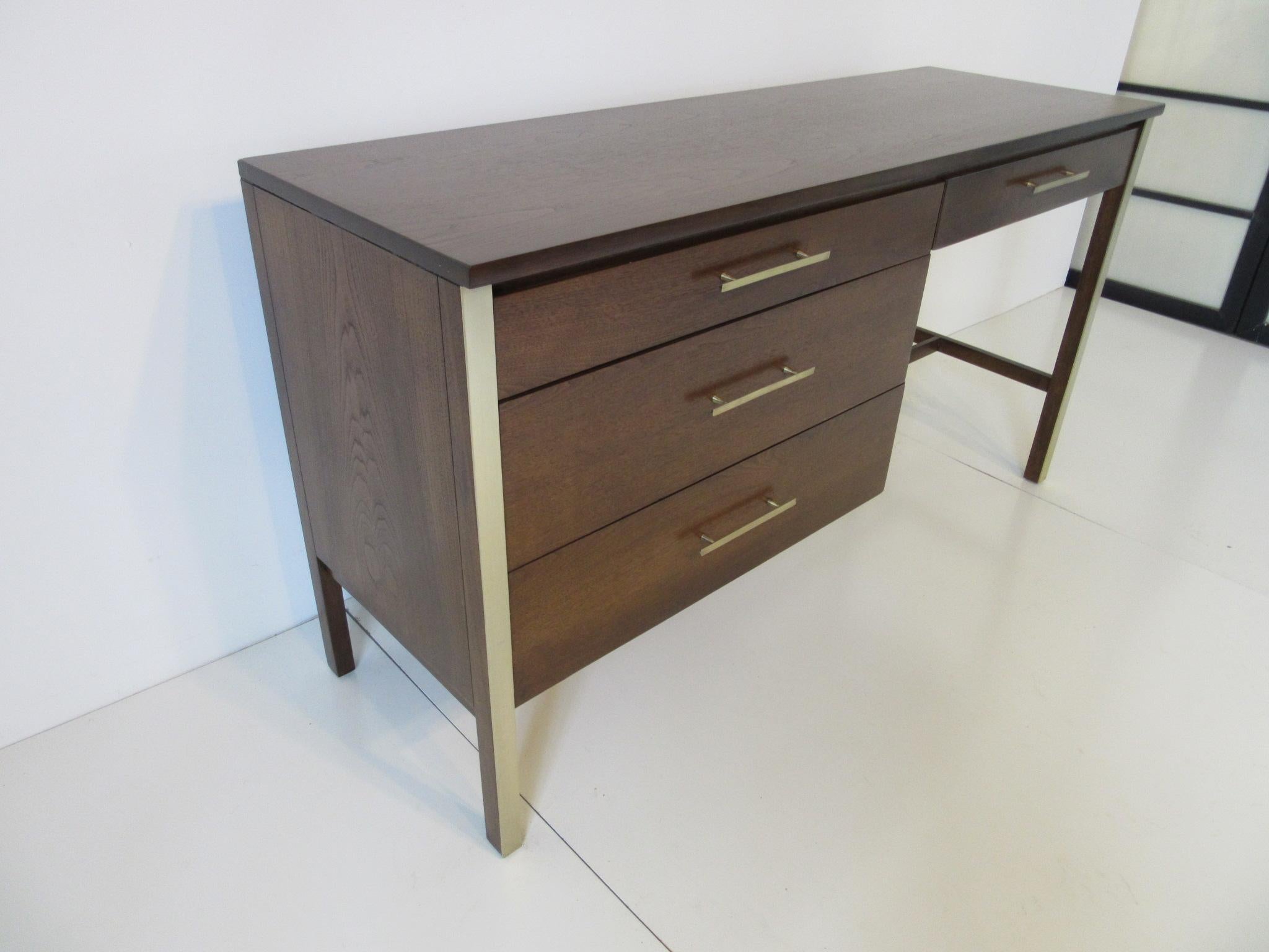 A dark walnut McCobb desk with four drawers and long brass pulls complemented with brass toned details to the front legs. A rare narrow styled desk which works well and takes up very little space.
  