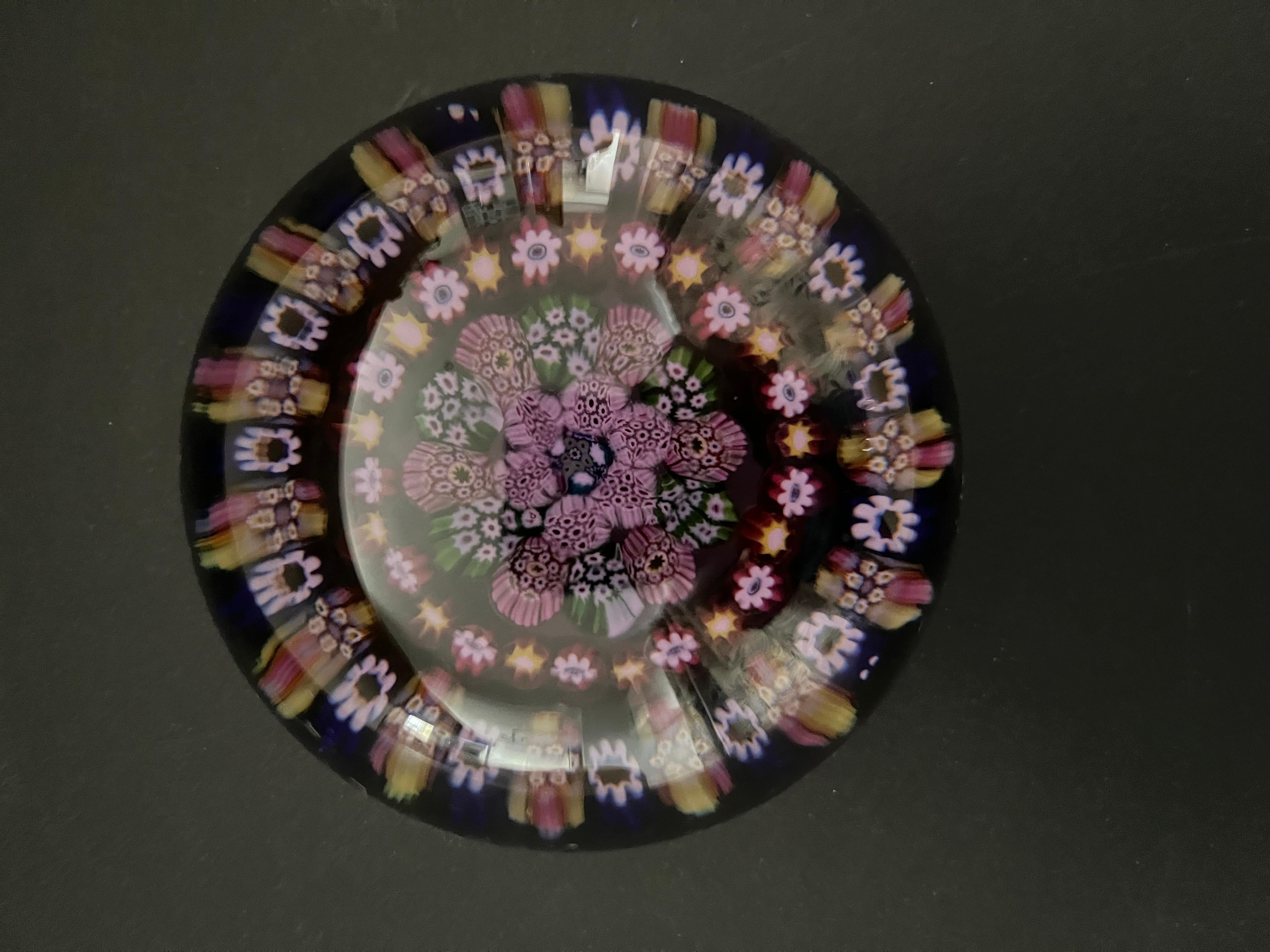 Si tratta di un bellissimo fermacarte in vetro scuro Millefiori concentrico fatto a mano, realizzato da Paul Ysart in Scozia, durante il suo primo periodo, circa negli anni '30.

Le canne sono disposte come due anelli concentrici aperti attorno a