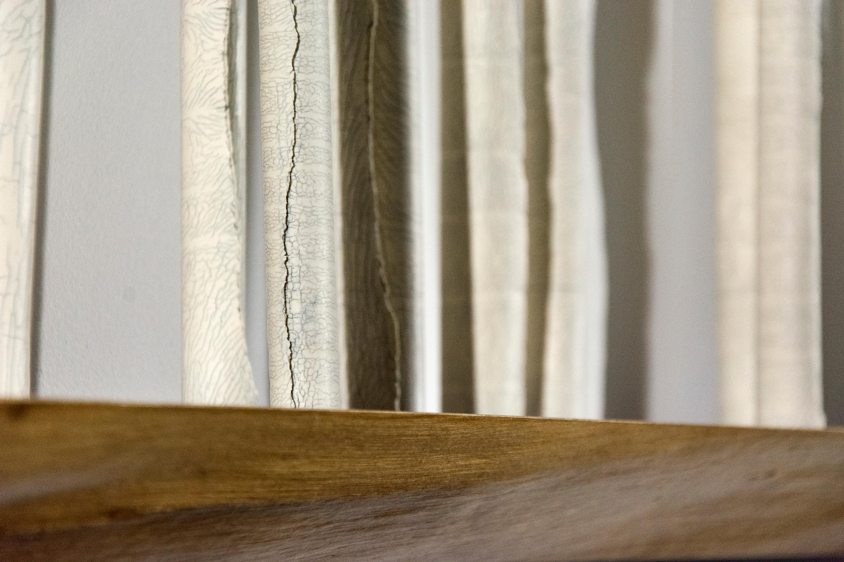 Porcelain scrolls stand on a walnut shelf in this poetic installation by Paula Murray.  The scrolls are articulated with fissures created through interaction of fiberglass filaments inserted into the wet porcelain clay before firing. Titled