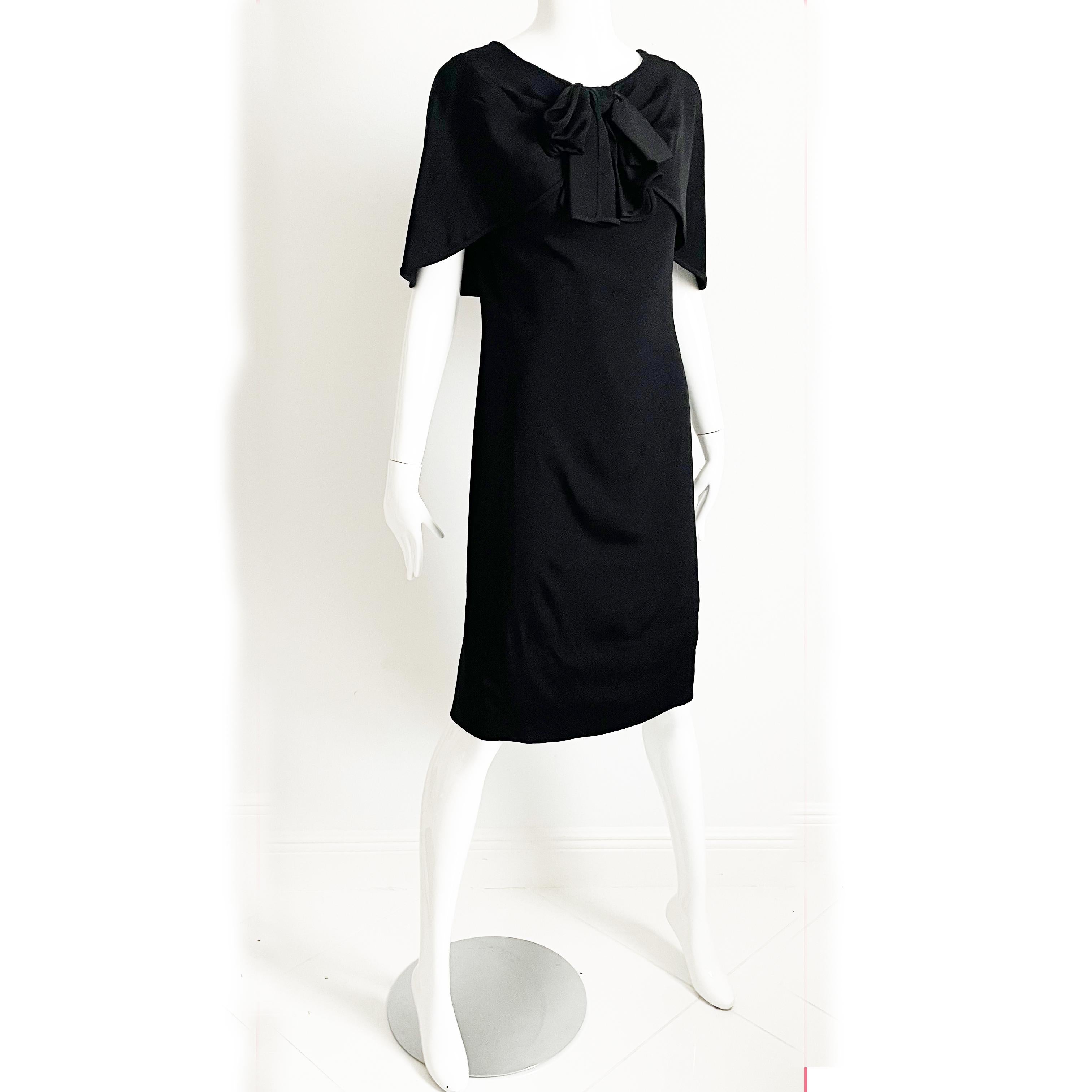 This incredible little black dress is an early Pauline Trigere piece, likely made in the late 1950s or early 60s.

Made from a supple black silk fabric, it features a draped shawl collar with a black silk bow design at the chest and a chic lower cut