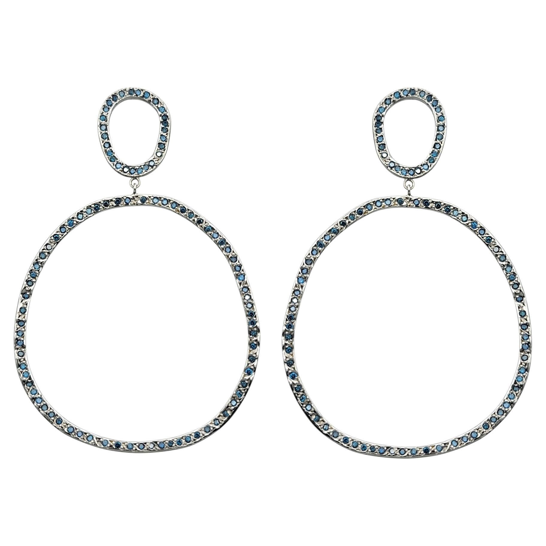 Diese Ohrringe aus 14-karätigem Weißgold mit doppeltem, offenem Kreis bestechen durch ihr modernes, luftiges Design, das vollständig von blau schimmernden Diamanten eingerahmt wird. Der obere Kreis geht nahtlos in einen größeren unteren Reif über,