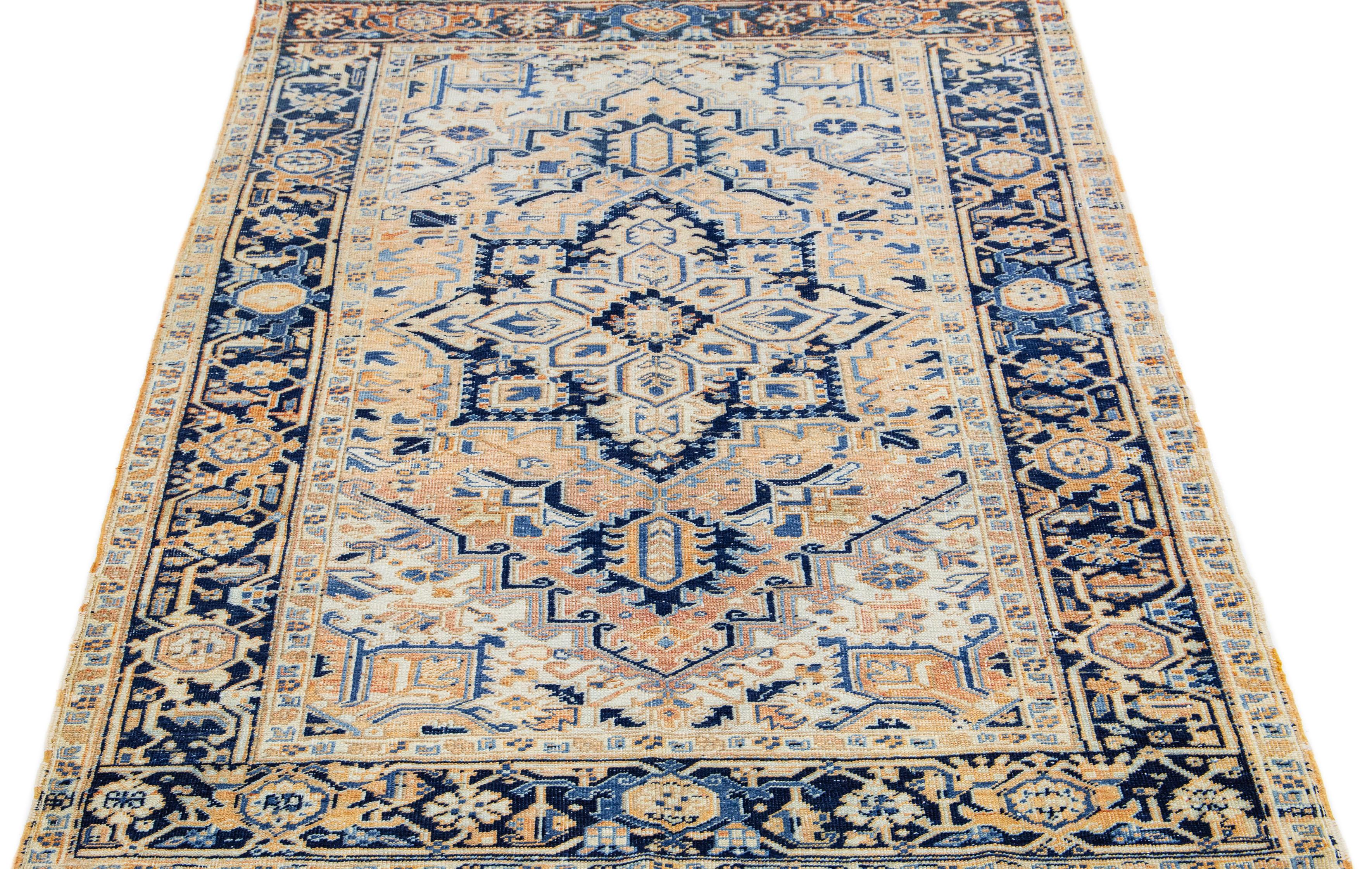 The antique Heriz rug's premium hand-knotted wool and striking medallion design exude timeless elegance and sophistication. The intricate geometric floral pattern in shades of blue and orange adds a touch of allure to the soft peach field, making it