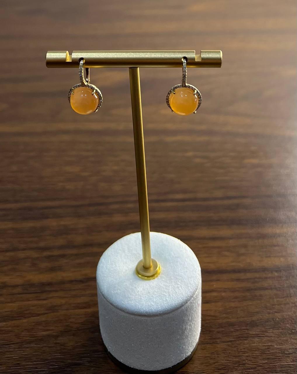 Un par de elegantes pendientes con gemas lunares redondas de color naranja melocotón, engastadas en oro de 14k y adornadas con delicados detalles de diamantes. El diseño está suspendido de una elegante bisagra dorada, que ofrece un aspecto refinado