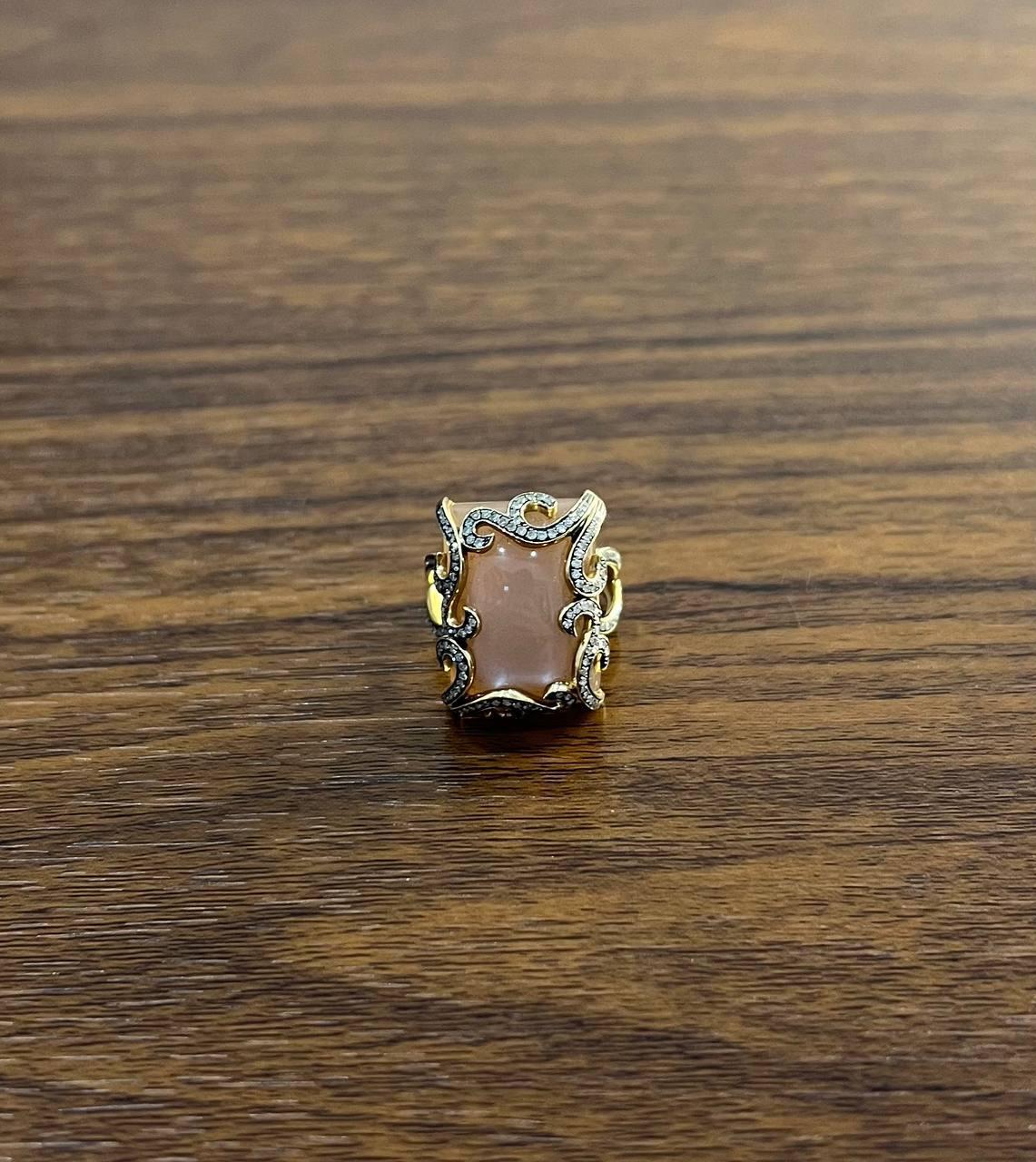 This exquisite ring features a captivating central peach pink moonstone with a soft, warm hue. The stone is elegantly framed by intricate scrollwork, adorned with delicate accent diamonds that enhance its allure.
Crafted in 14k gold.
Primary Carat
