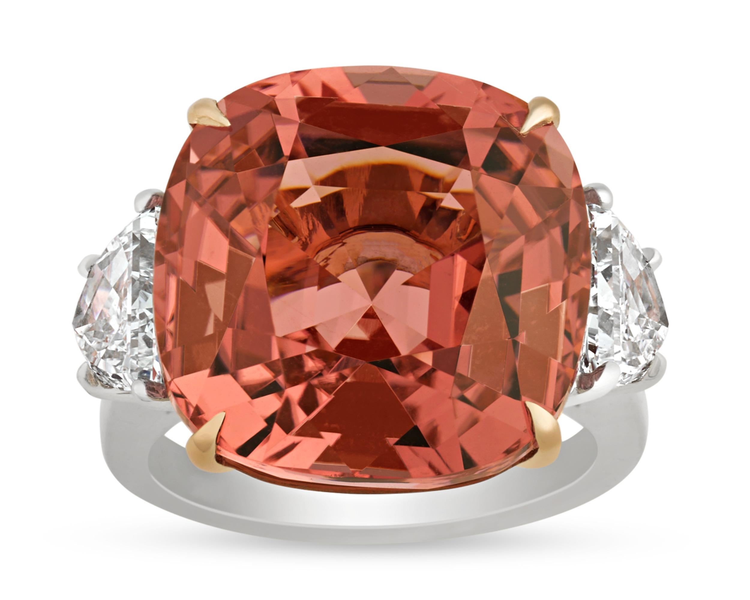 A stunning cushion-cut tourmaline weighing 19.74 carats displays a unique peachy hue at the center of this classic ring. The gemstone's romantic, orangy-pink color is enhanced by two half-moon diamonds weighing a total of 1.53 carats that display G