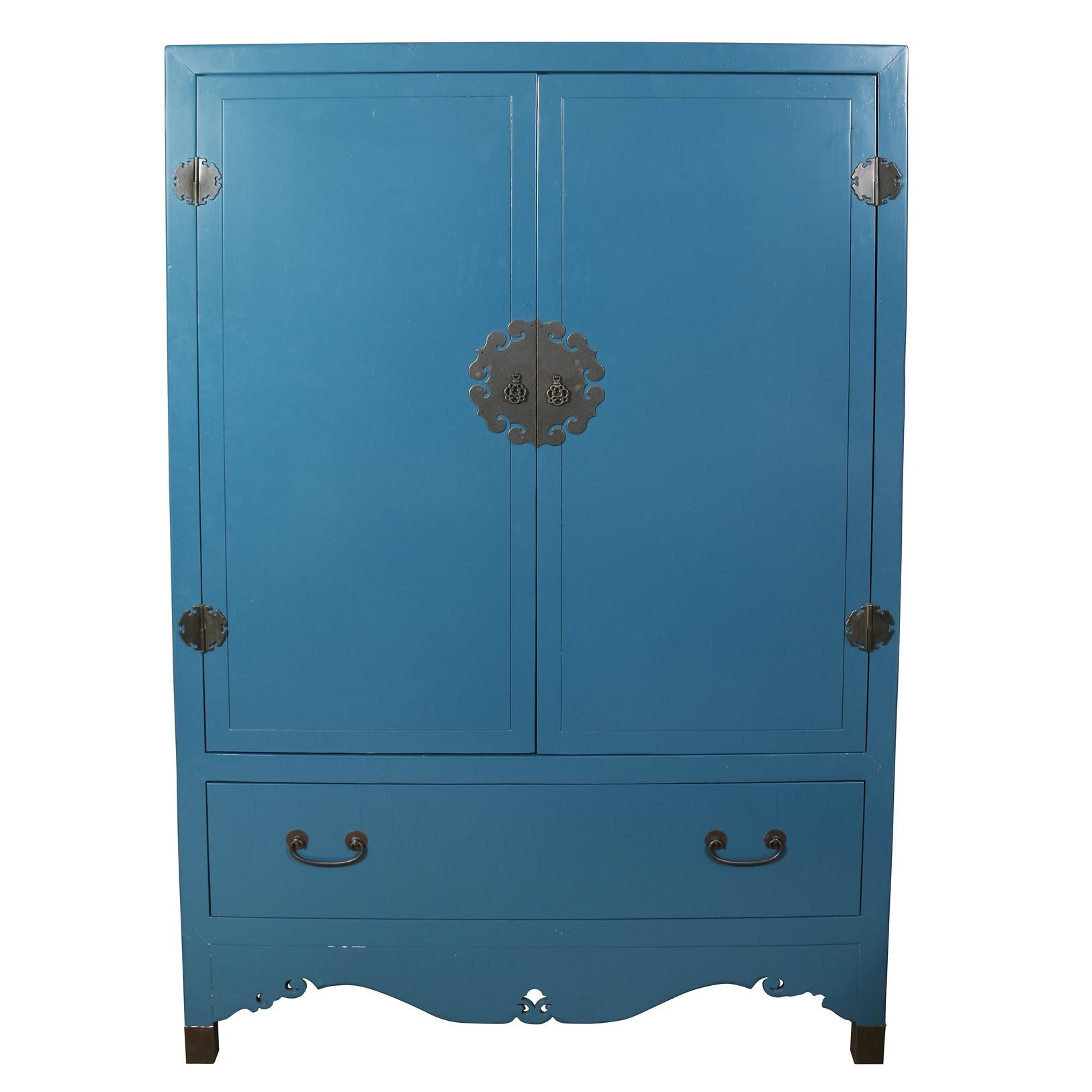 A peacock blue Asian style armoire with brass fittings. A round brass medallion seen in Asian decor surrounds the door hardware for the center door pulls. The beautiful blue armoire opens to reveal a large interior with openings for electrical