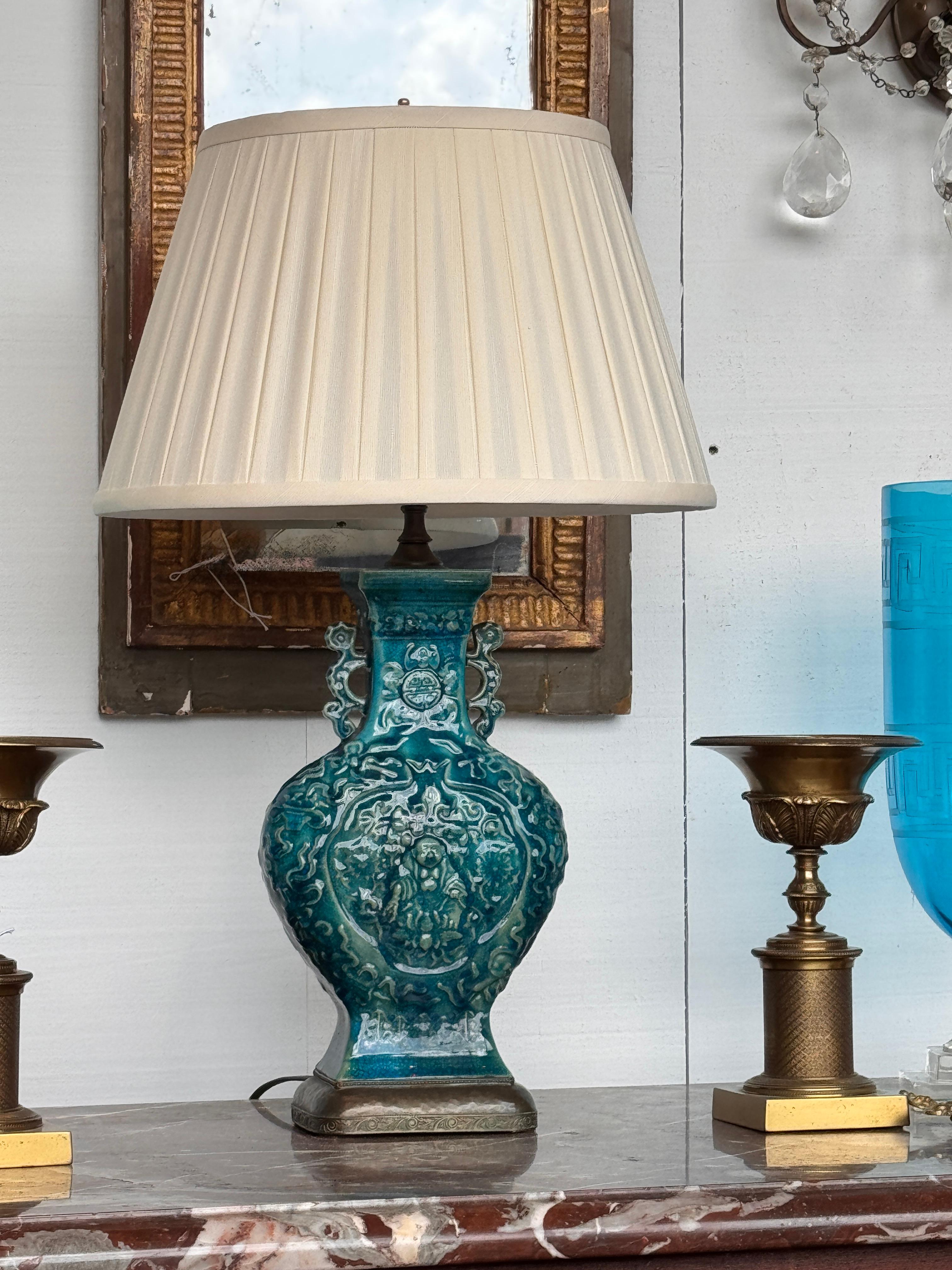 Chinese ceramic lamp in a deep peacock blue glaze, featuring molded figural and foliate relief work over a crackled surface. The vase-form body is mounted on an embossed bronze base with a hammered finish and engraved detailing along the border.
