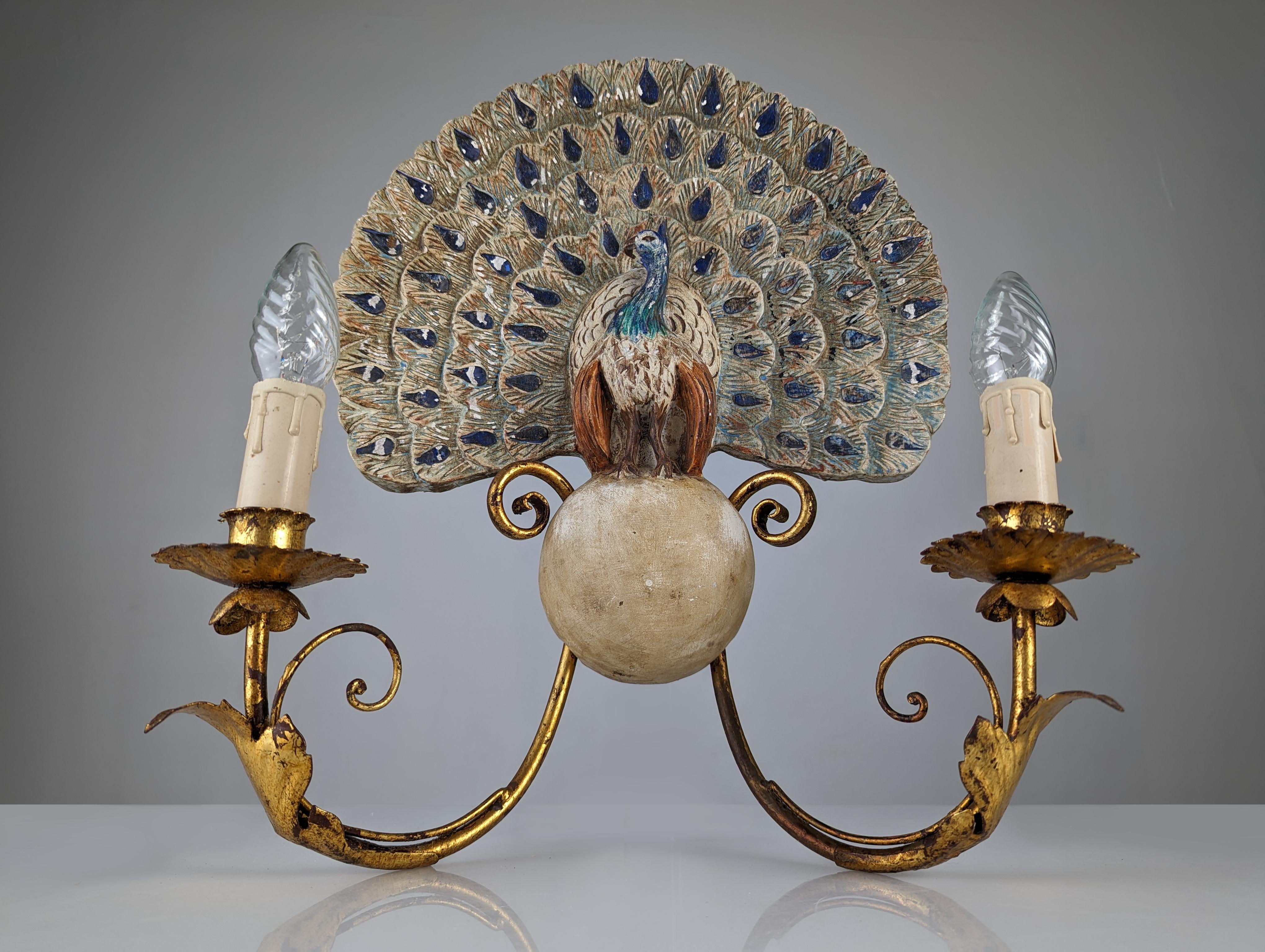 Special peacock applique with its beautiful extended tail carved in polychrome wood perched on a wooden sphere from which two arms hang, decorated with golden foliage. A fantastic work from the Early 20th Century.