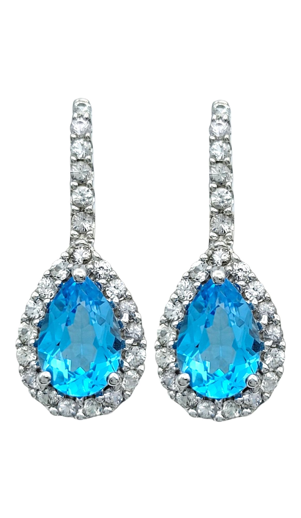 These exquisite drop earrings dazzle with vibrant pear-shaped blue topaz gemstones, their luminous oceanic hue radiating elegance and charm. Encased in a sparkling halo of brilliant white sapphires, the design enhances the depth and brilliance of