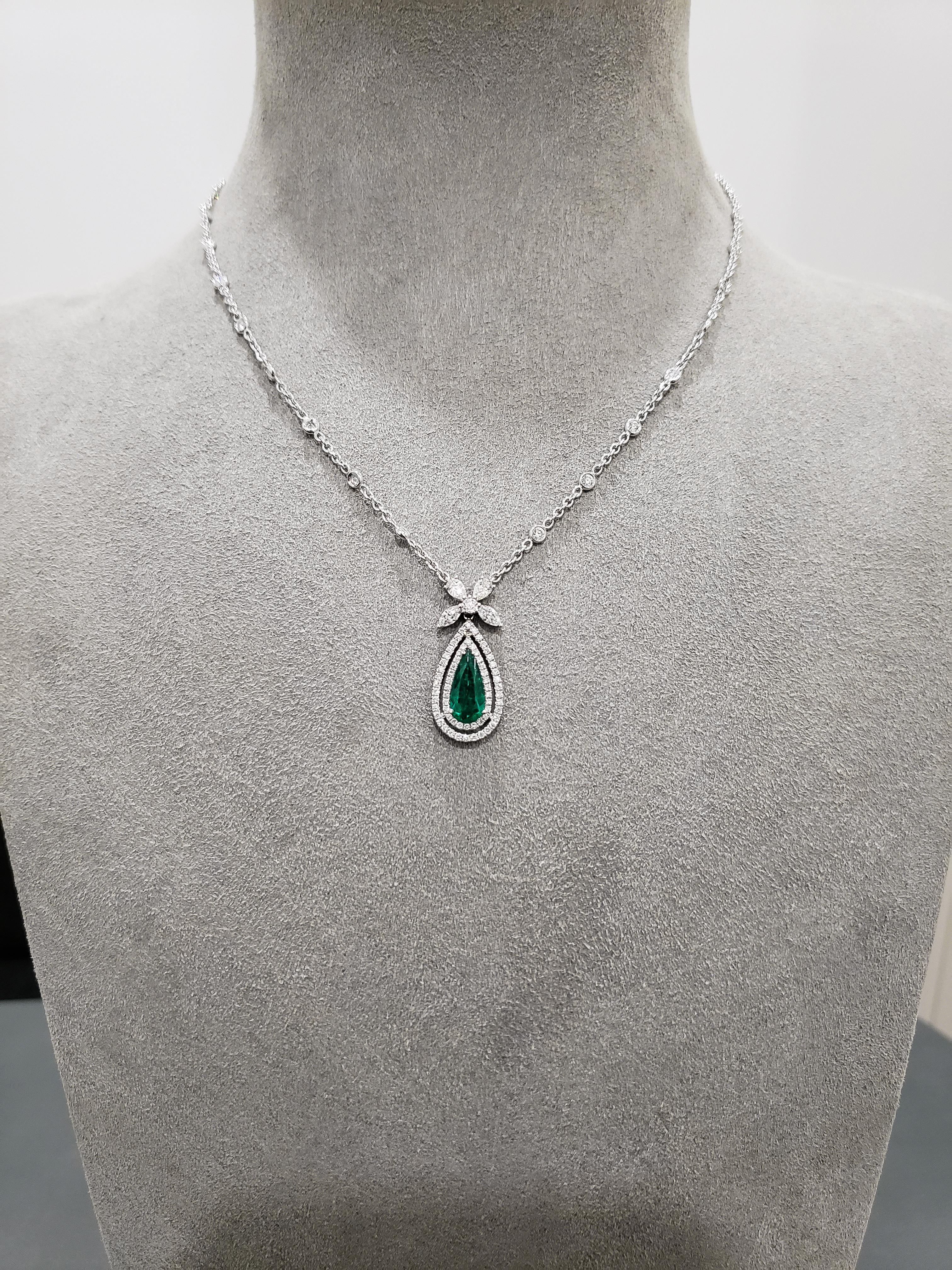 A color-rich pendant necklace showcasing a 1.01 carat pear-shaped green emerald, accented by two rows of round brilliant diamonds in an open-work design. Attached to a bow-like figure made with diamonds. Suspended on a diamonds by the yard chain.