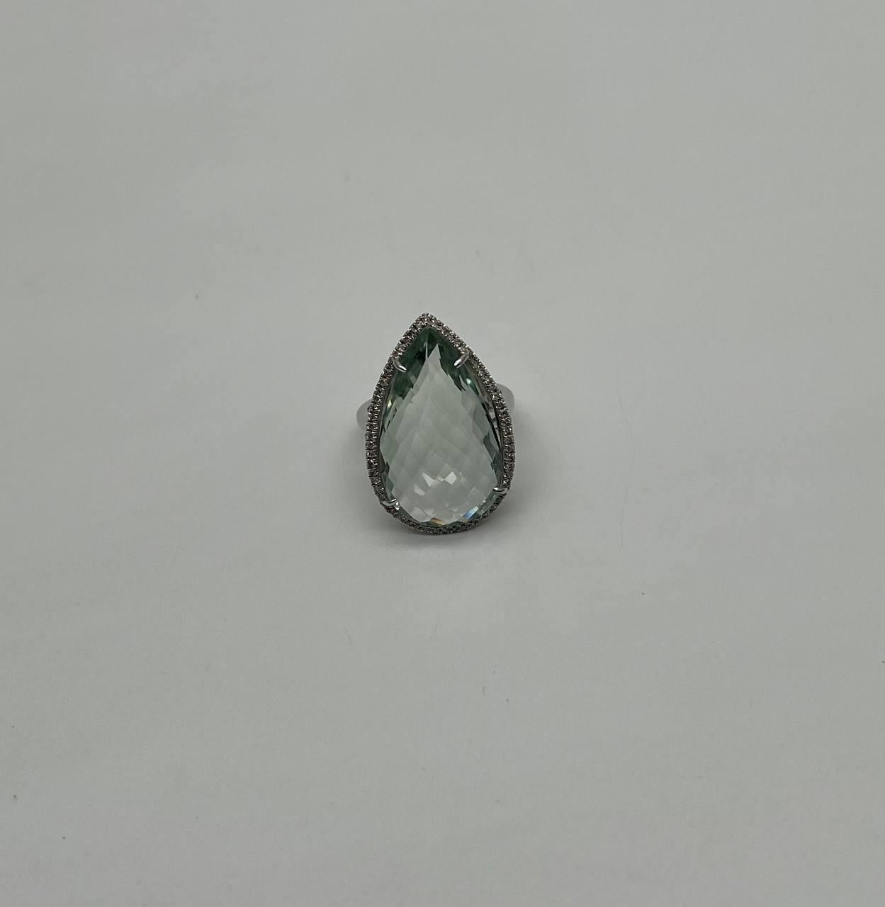 This statement ring is crafted in 14k white gold.  It features a large, faceted teardrop-shaped blue topaz, surrounded by a delicate halo of small, sparkling diamonds. The setting is sleek and elegant, enhancing the brilliance of the central gem.