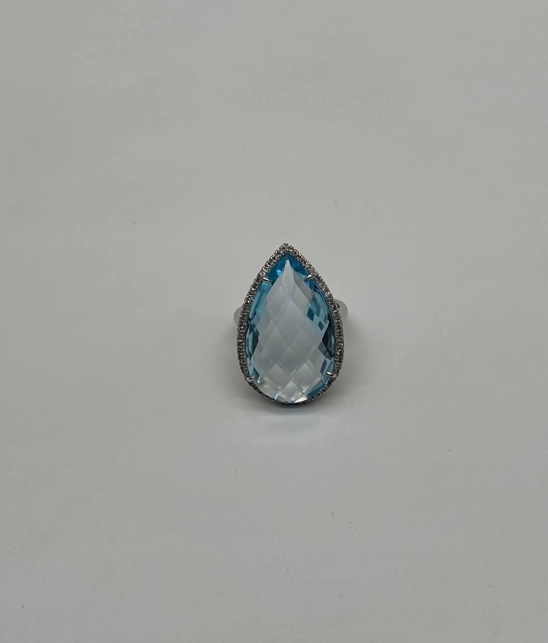 A striking ring featuring a large, faceted blue topaz set in a detailed 14K White Gold band. The gemstone is pear-shaped, with intricate facets that catch the light beautifully. The white gold setting is adorned with a delicate border halo of white