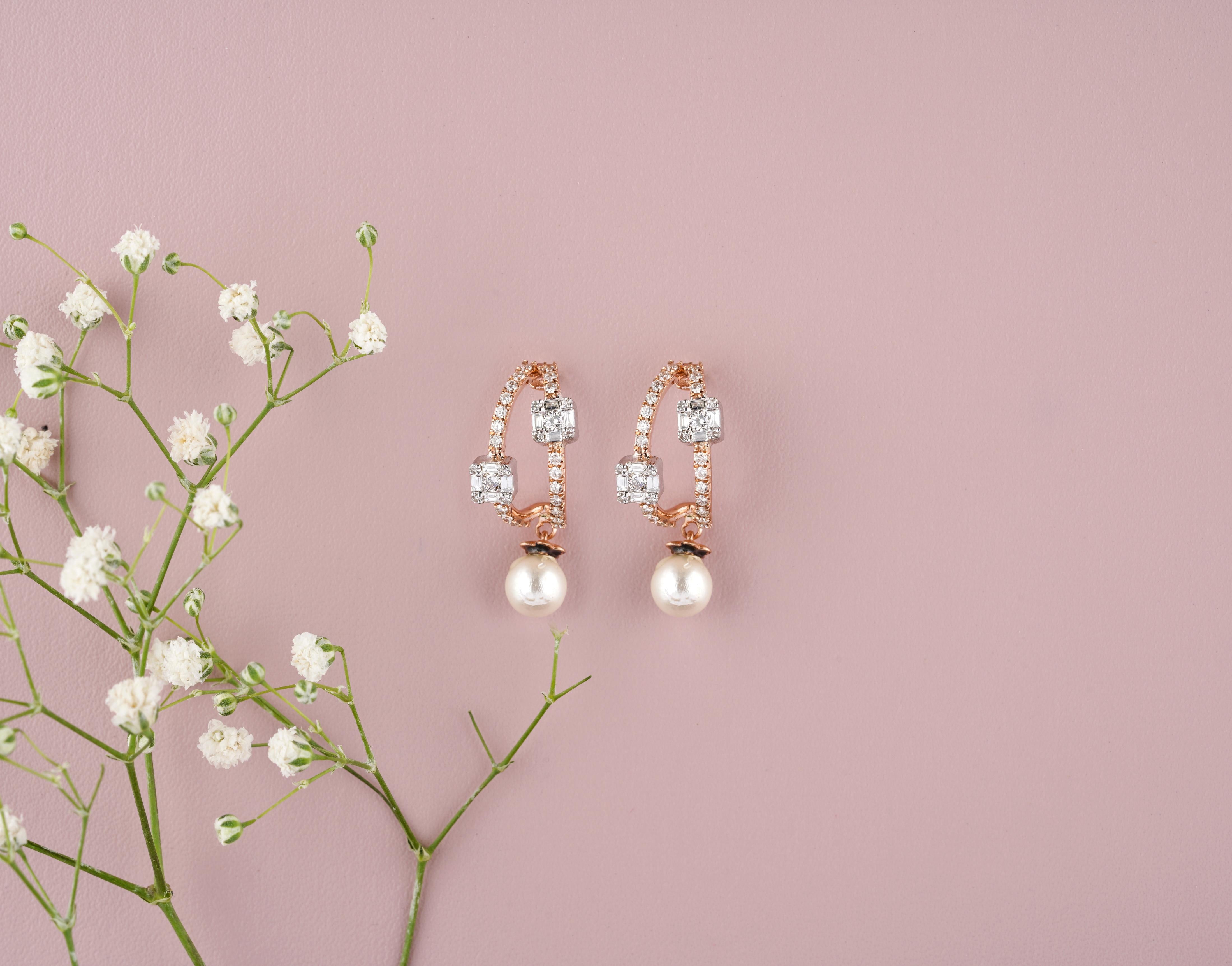 Pearl Drop Diamond Earrings feature a hoop design crafted in rose gold, each adorned with three emerald-cut diamonds evenly spaced along the front of the hoop. Dangling gracefully from the bottom of each hoop is a single lustrous pearl, adding an